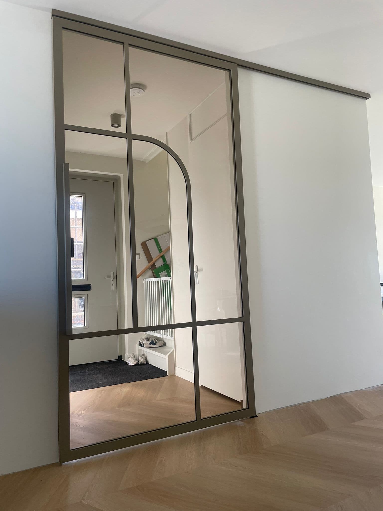 Gesloten bronzen stalen schuifdeur met een subtiel boogdesign en brons getint glas, schuivend langs een witte wand.