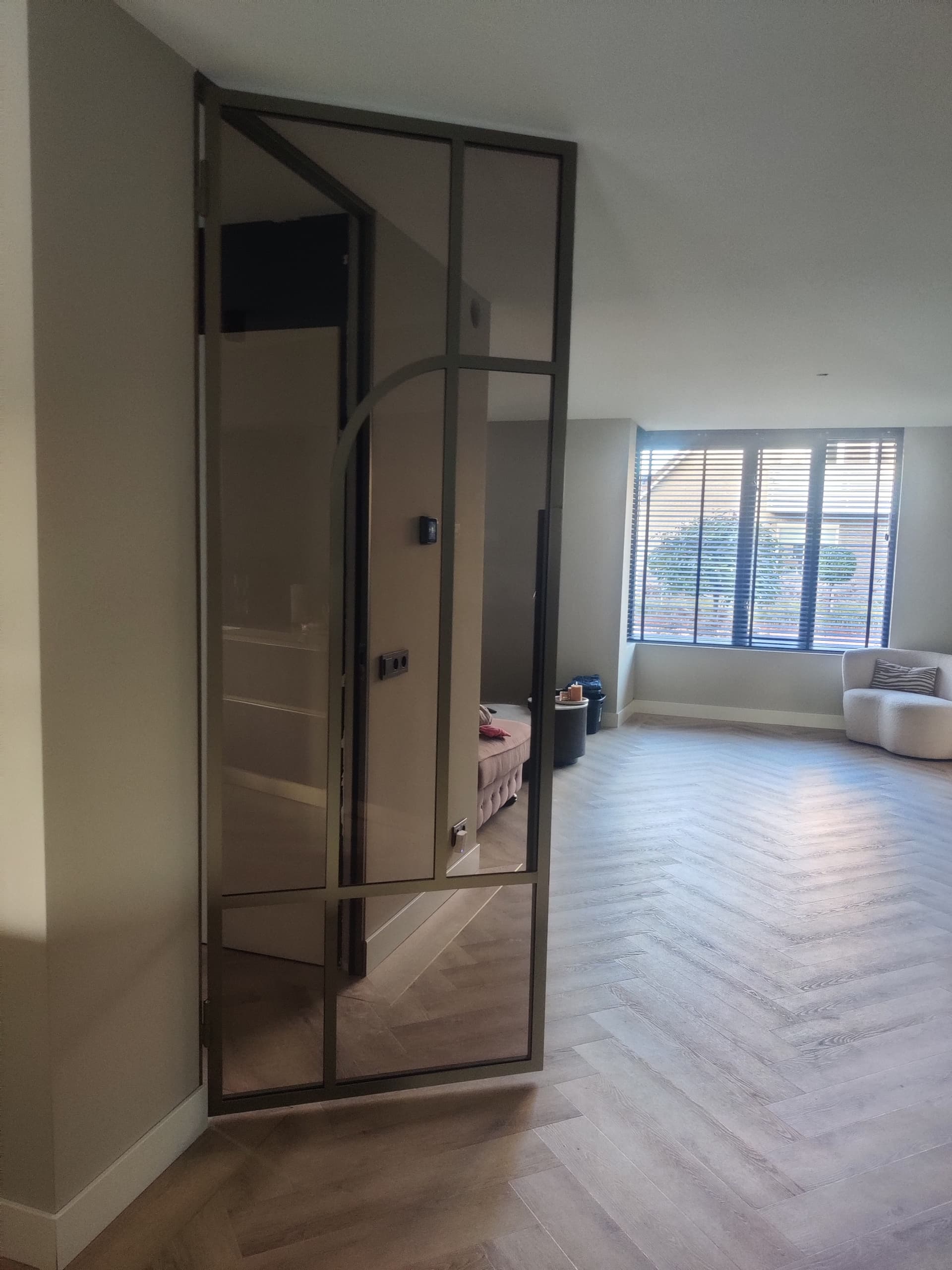 Kamerhoge bruine stalen scharnierdeur met een subtiel boogdesign en brons getint glas in een interieur met visgraatvloer.