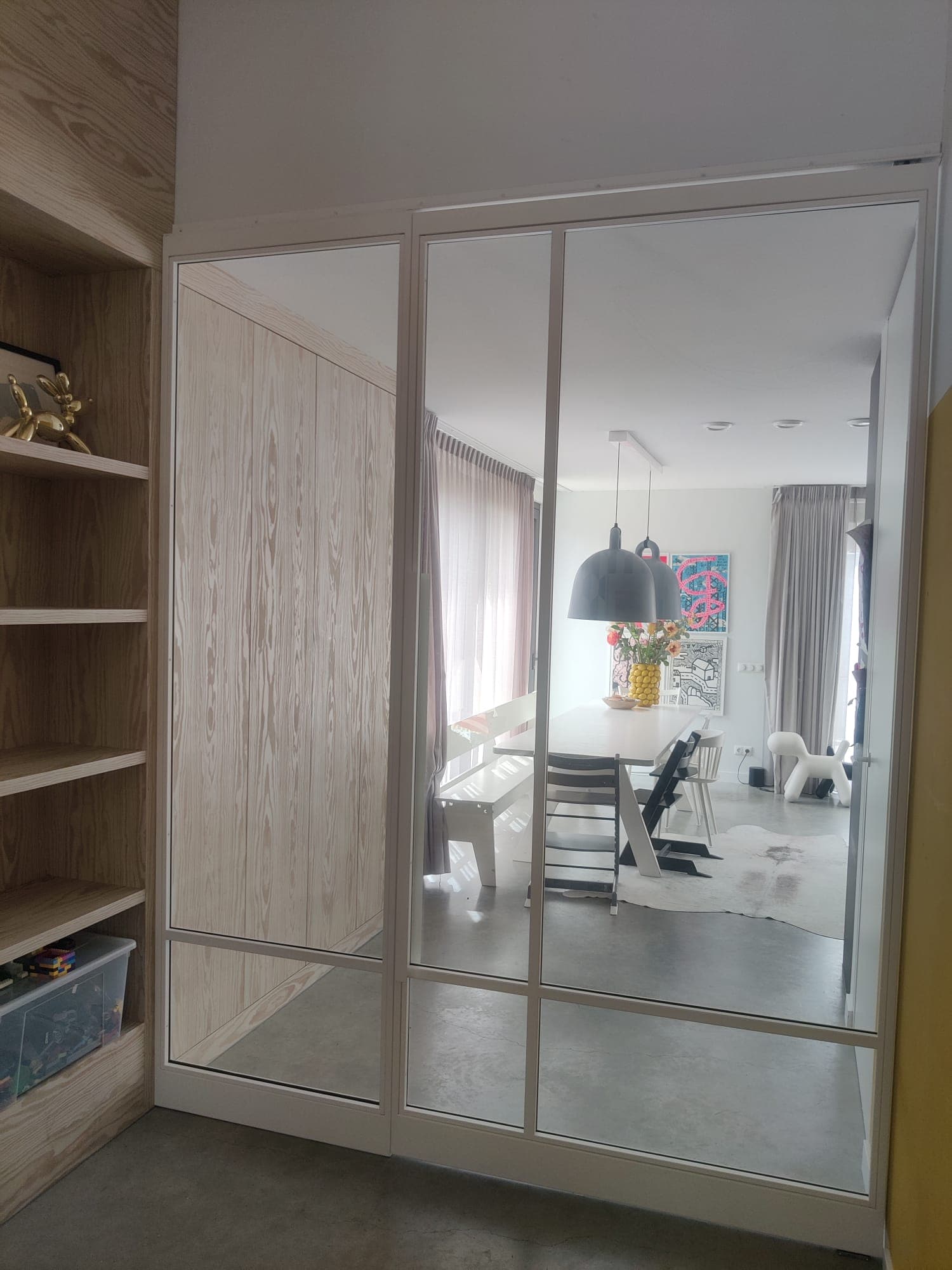 Brede witte stalen taatsdeur met vast zijpaneel tussen een hal en een eetkamer met hanglampen.