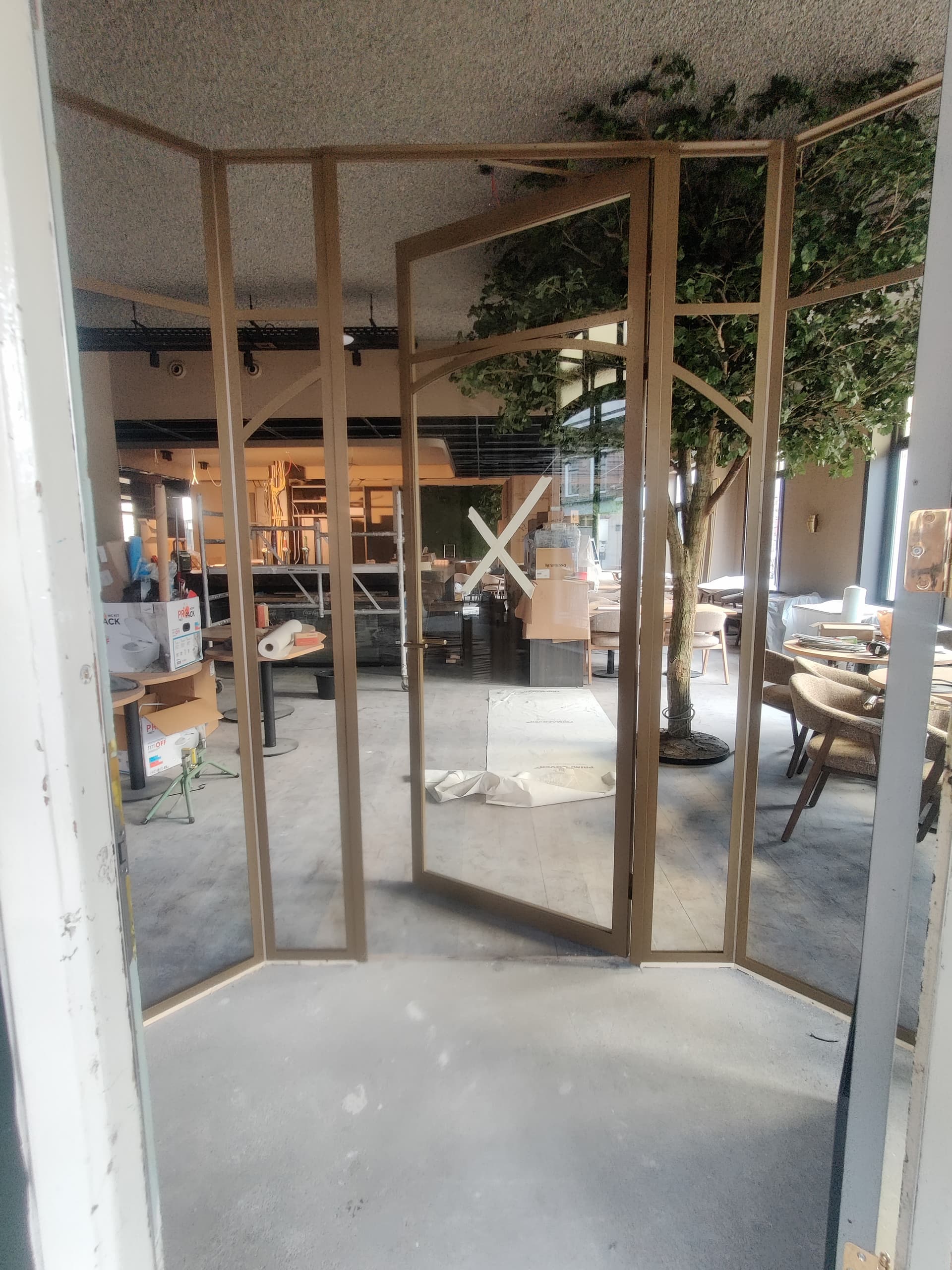 Op maat gemaakte goudkleurige stalen tochtsluis met een geopende binnendeur en helder glas in het interieur van een sfeervol café.