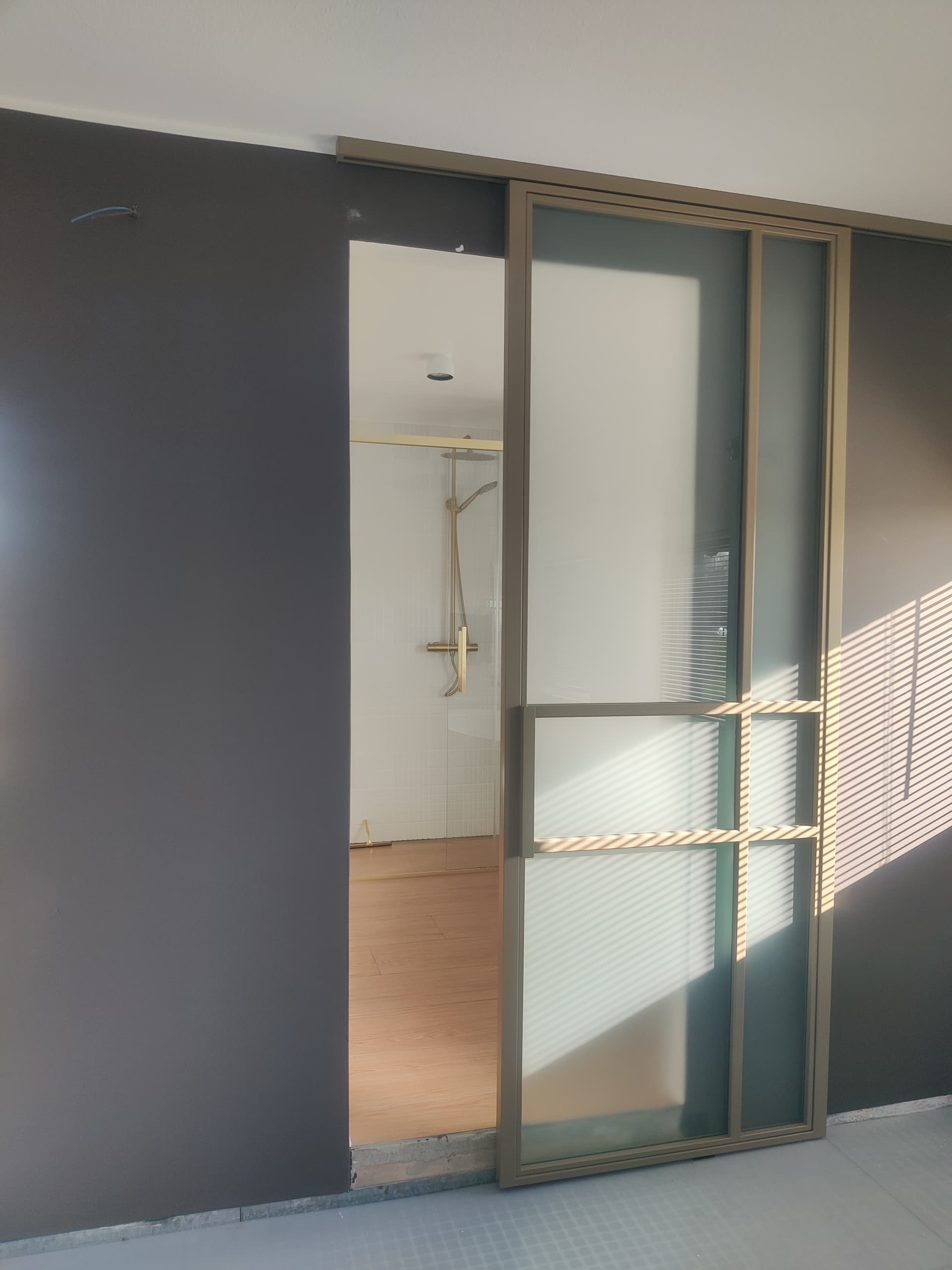 Geopende goud-bronskleurige stalen schuifdeur met frosted glas, met zicht op een moderne badkamer met gouden doucheset.