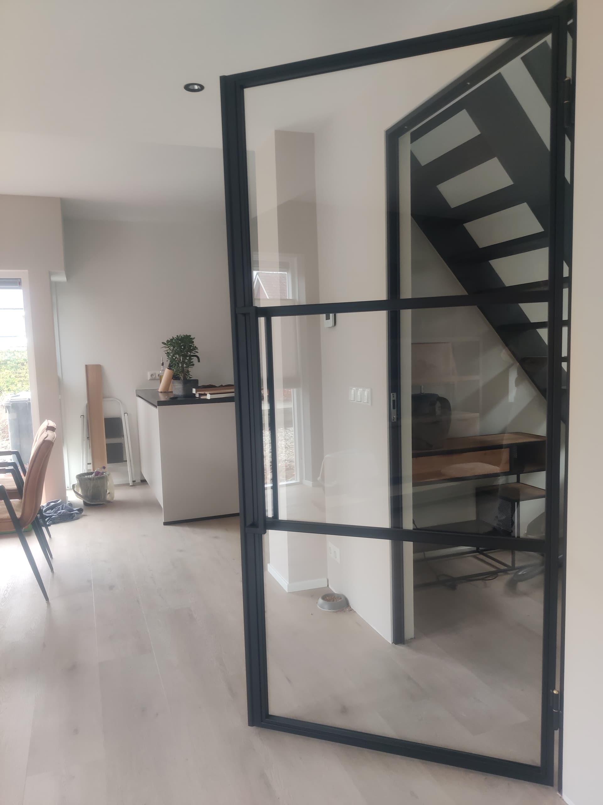 Lichte woonkamer met een openstaande zwarte stalen deur met helder veiligheidsglas en een strakke vakverdeling.