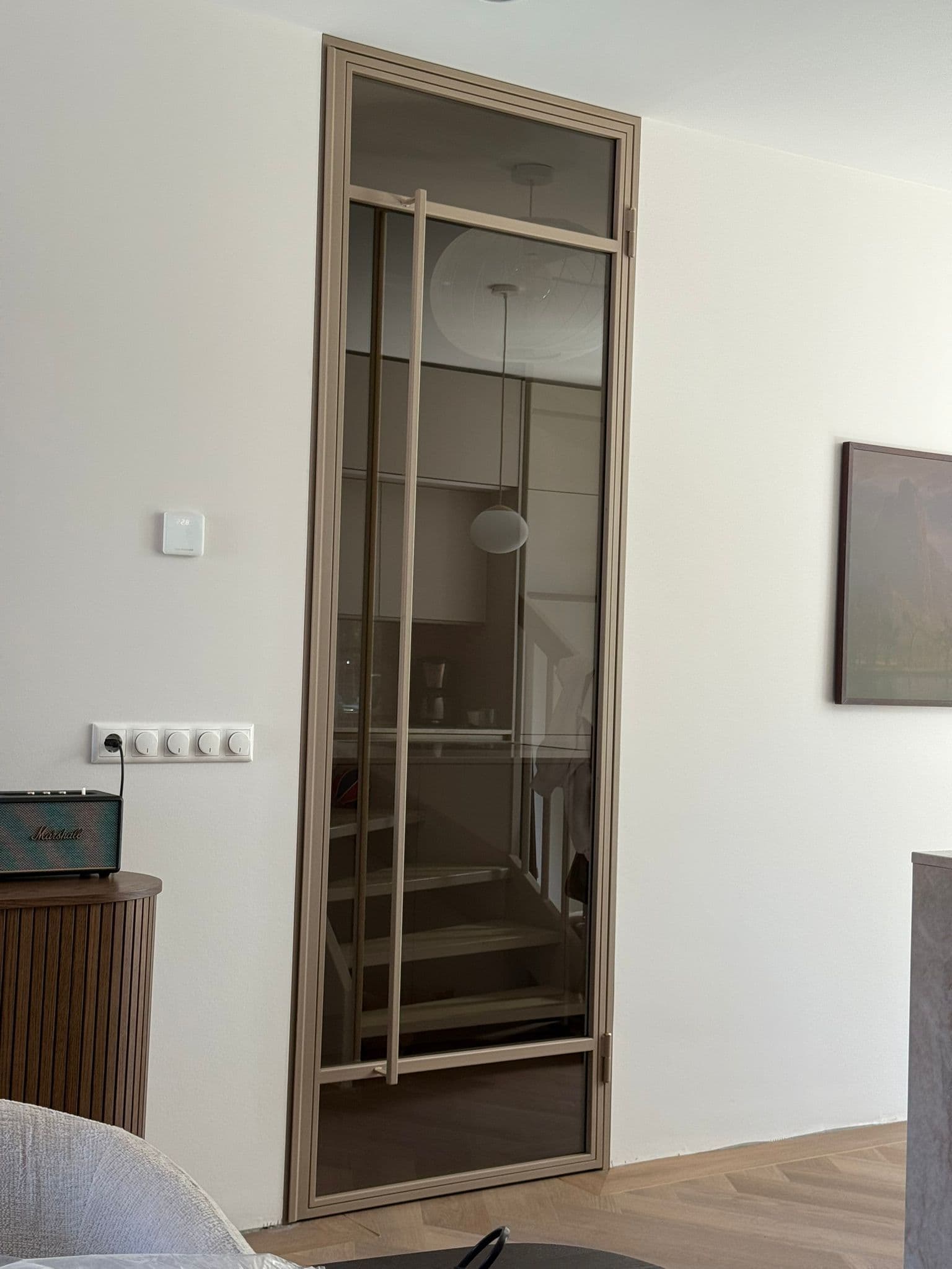 Elegante bronzen stalen scharnierdeur in kozijn met brons getint glas en een lange verticale handgreep in een modern interieur met visgraatvloer