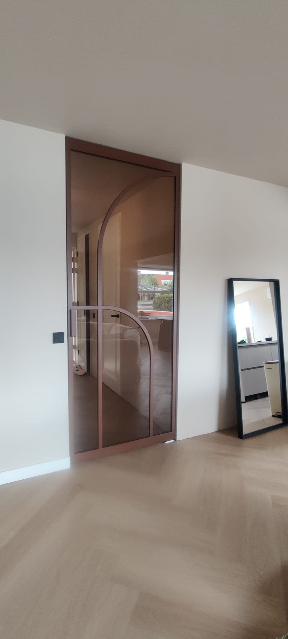 Enkele bronzen stalen deur met een asymmetrisch boogontwerp en brons getint glas in een modern interieur met visgraatvloer.