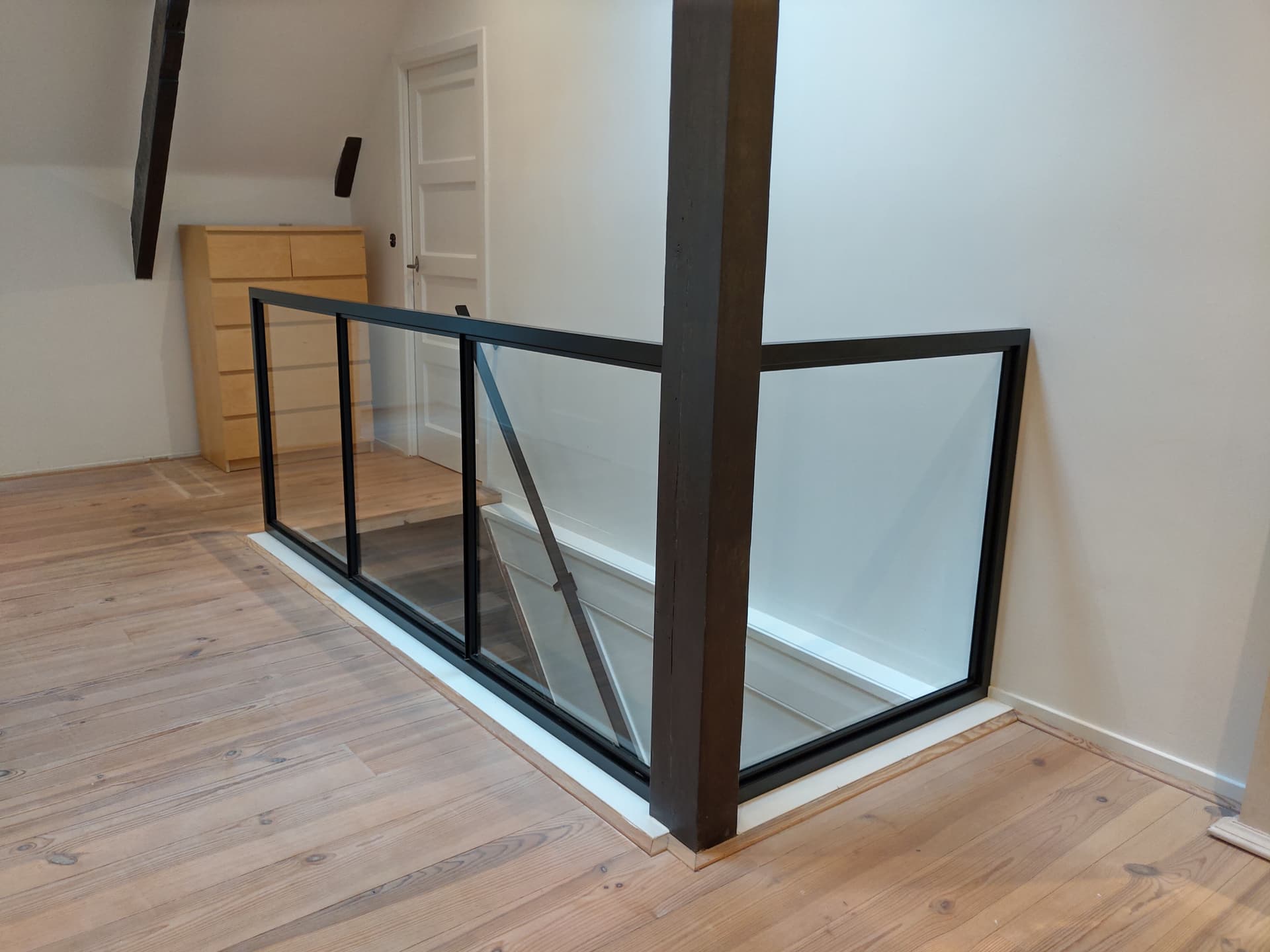 Zwarte stalen balustrade met glas rond een trapgat op een zolderverdieping met houten vloer.