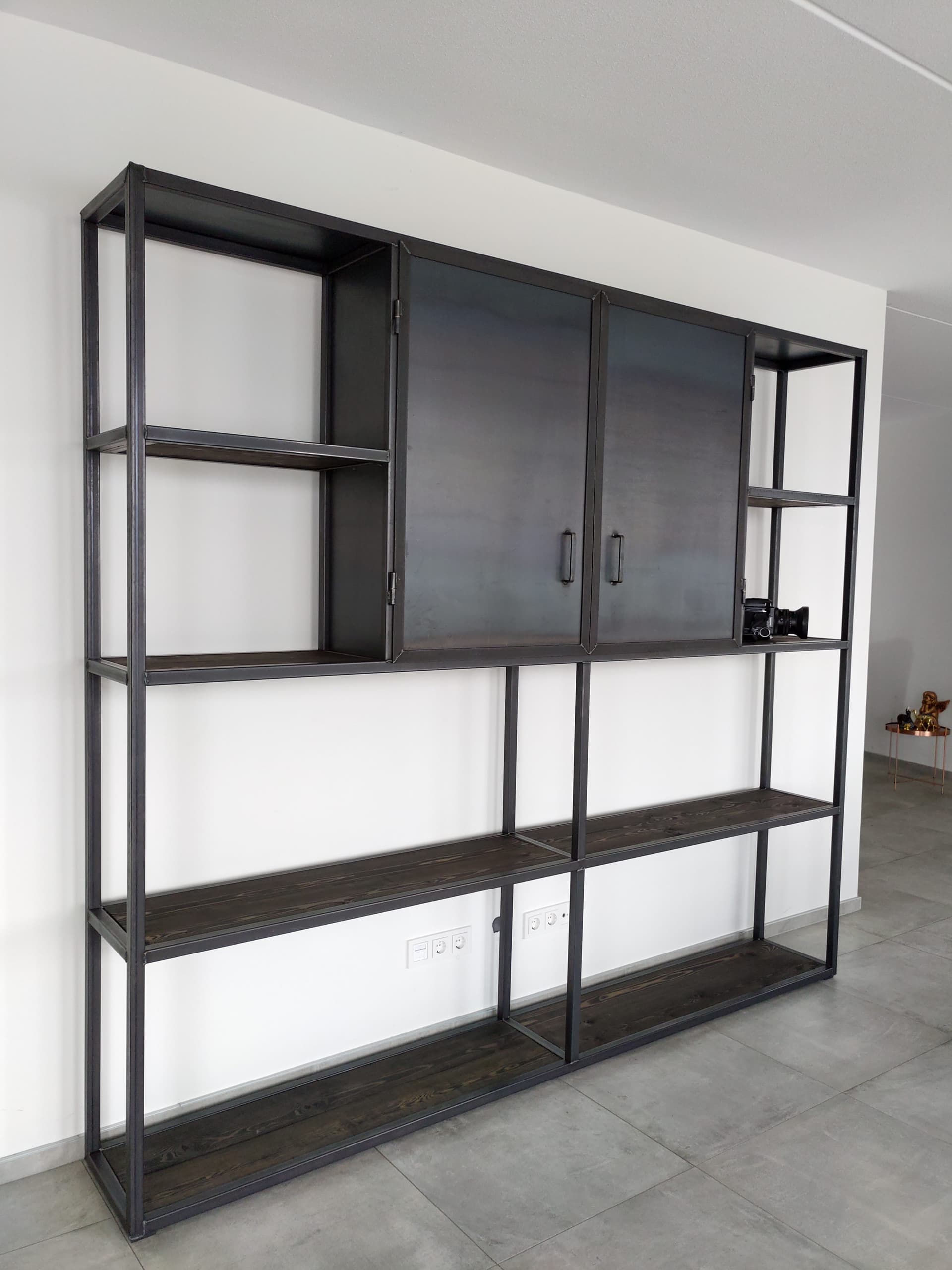 Vrijstaande stalen stellingkast met open vakken, houten planken en twee dichte kastdeuren van ruw staal tegen een witte wand.