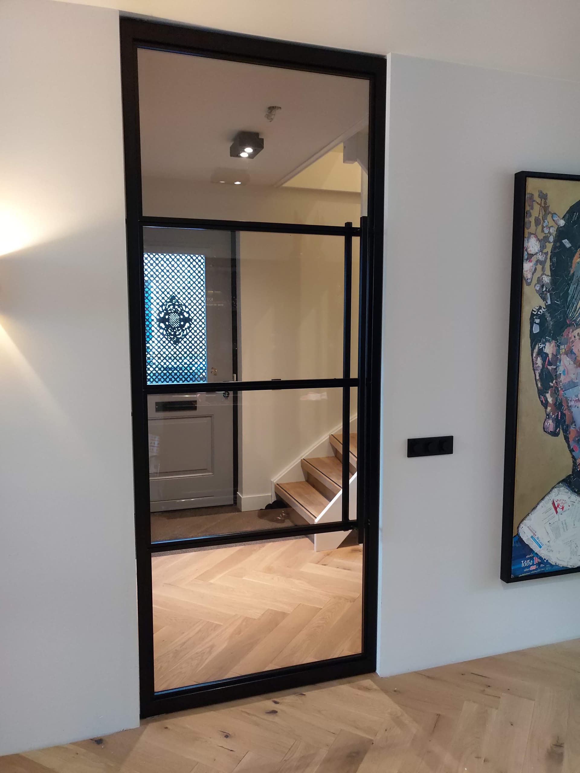 Enkele zwarte stalen taatsdeur met getint glas en een asymmetrische vakverdeling, met zicht op een hal met visgraatvloer.