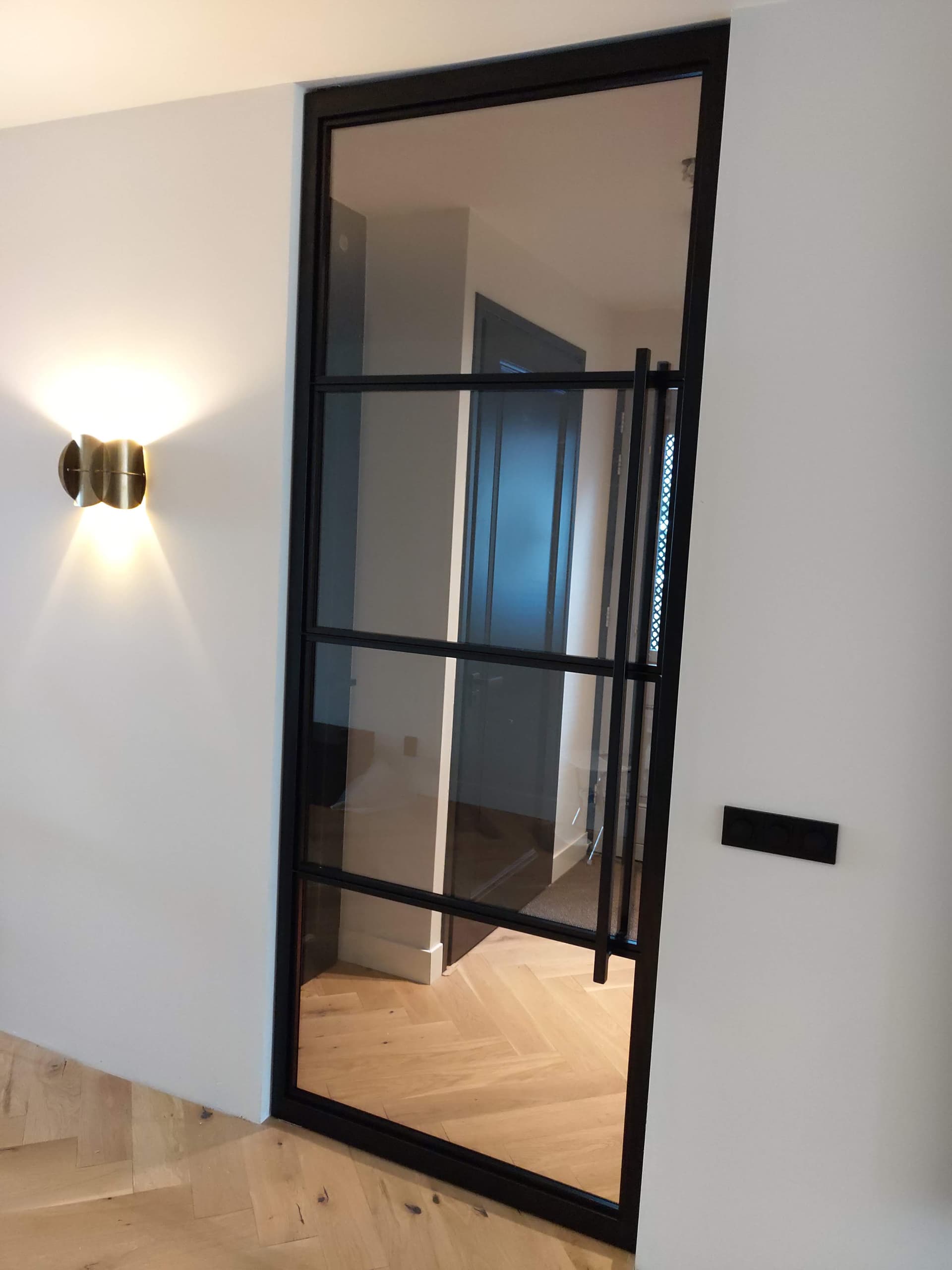 Gesloten zwarte stalen taatsdeur met getint glas en een lange verticale greep, in een hal met visgraatvloer.