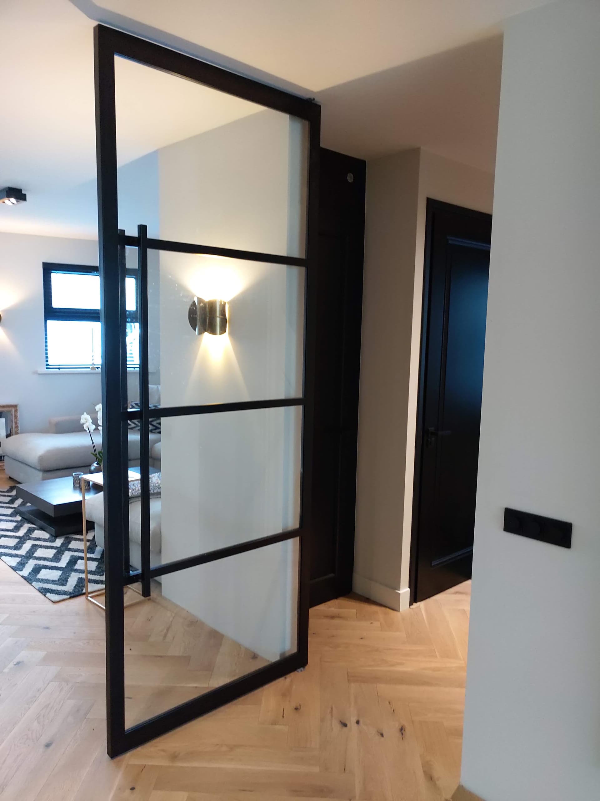 Openstaande zwarte stalen taatsdeur met deels matglas, met zicht op een woonkamer met wandlamp en visgraatvloer.