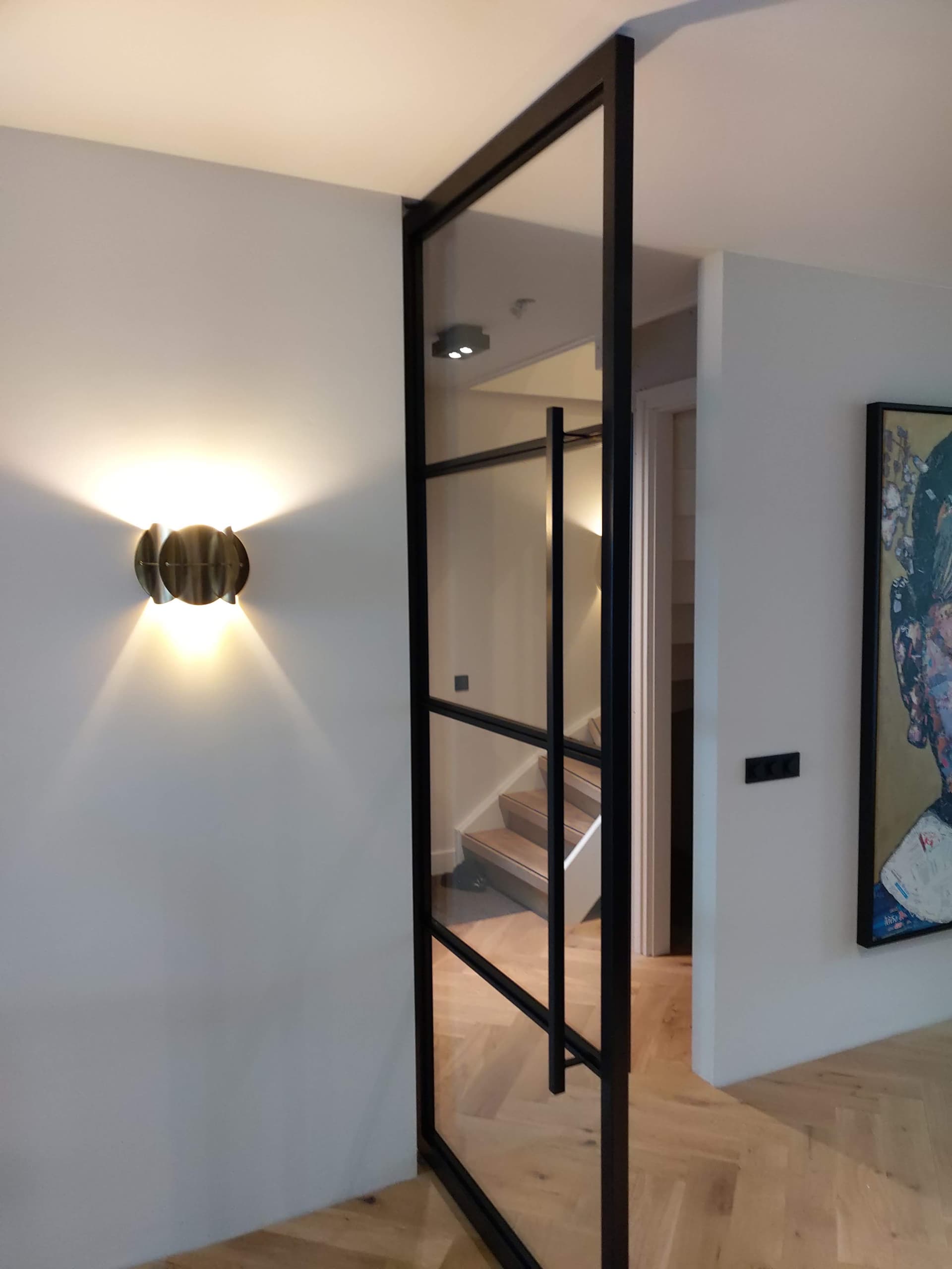 Zwarte stalen taatsdeur met getint glas half geopend in een hal, met zicht op een trap en een wandlamp.