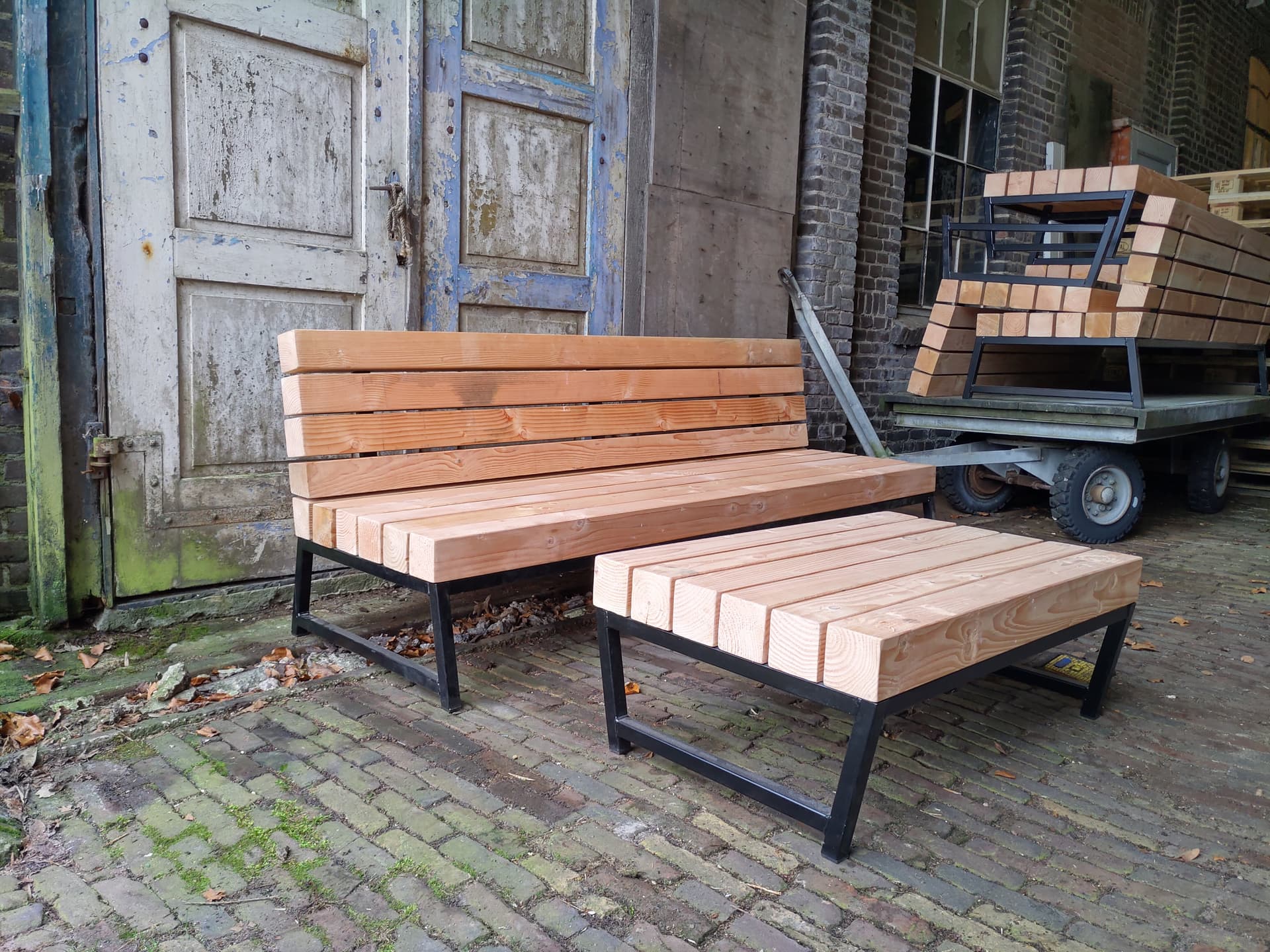 Tuinbank en bijpassende lage tafel van zwart staal met houten planken, op een klinkerbestrating voor een oude deur.
