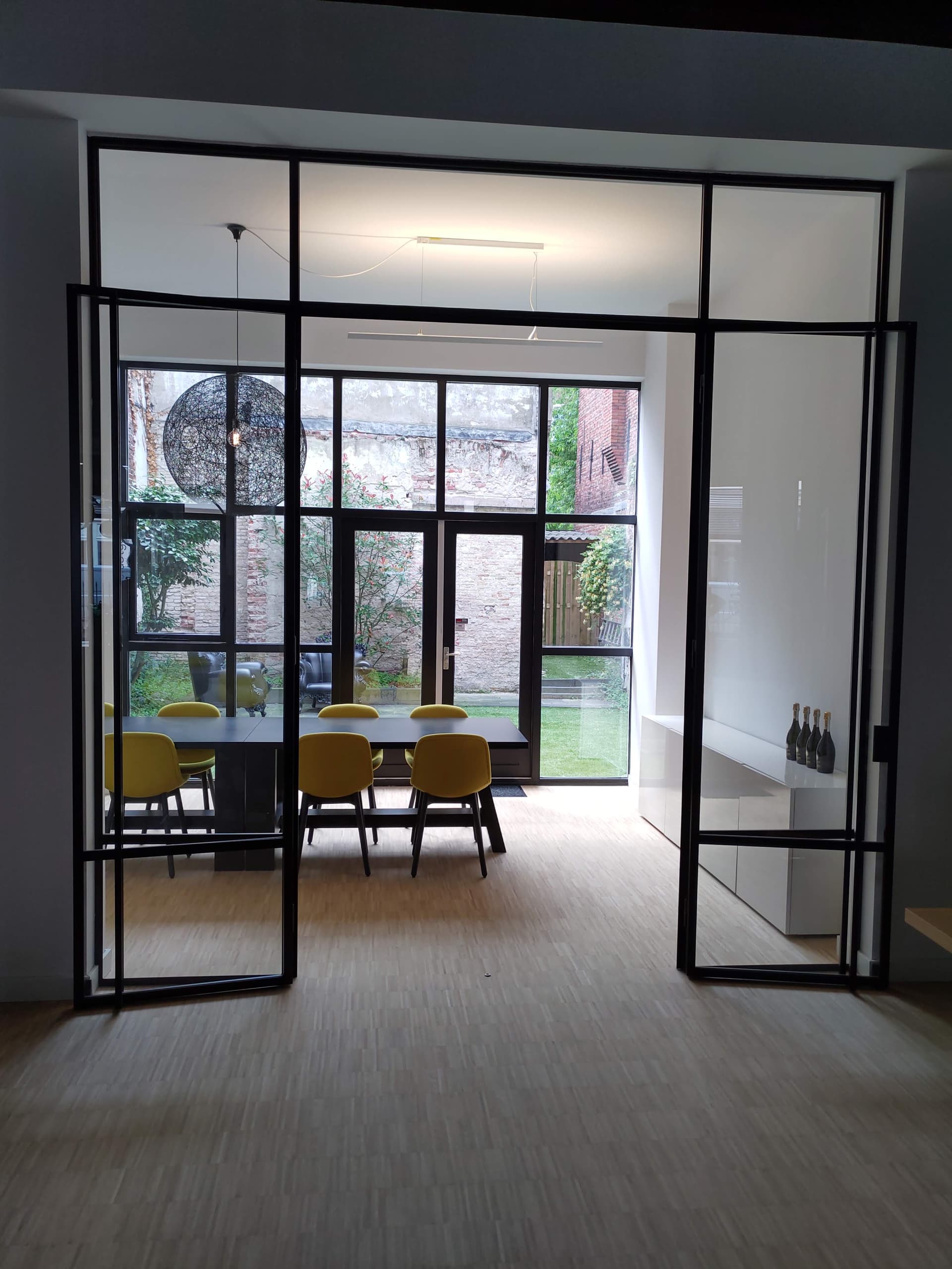 Openstaande zwarte stalen binnenpui met dubbele deuren en boven- en zijlichten, met doorkijk naar eetkamer en tuin.