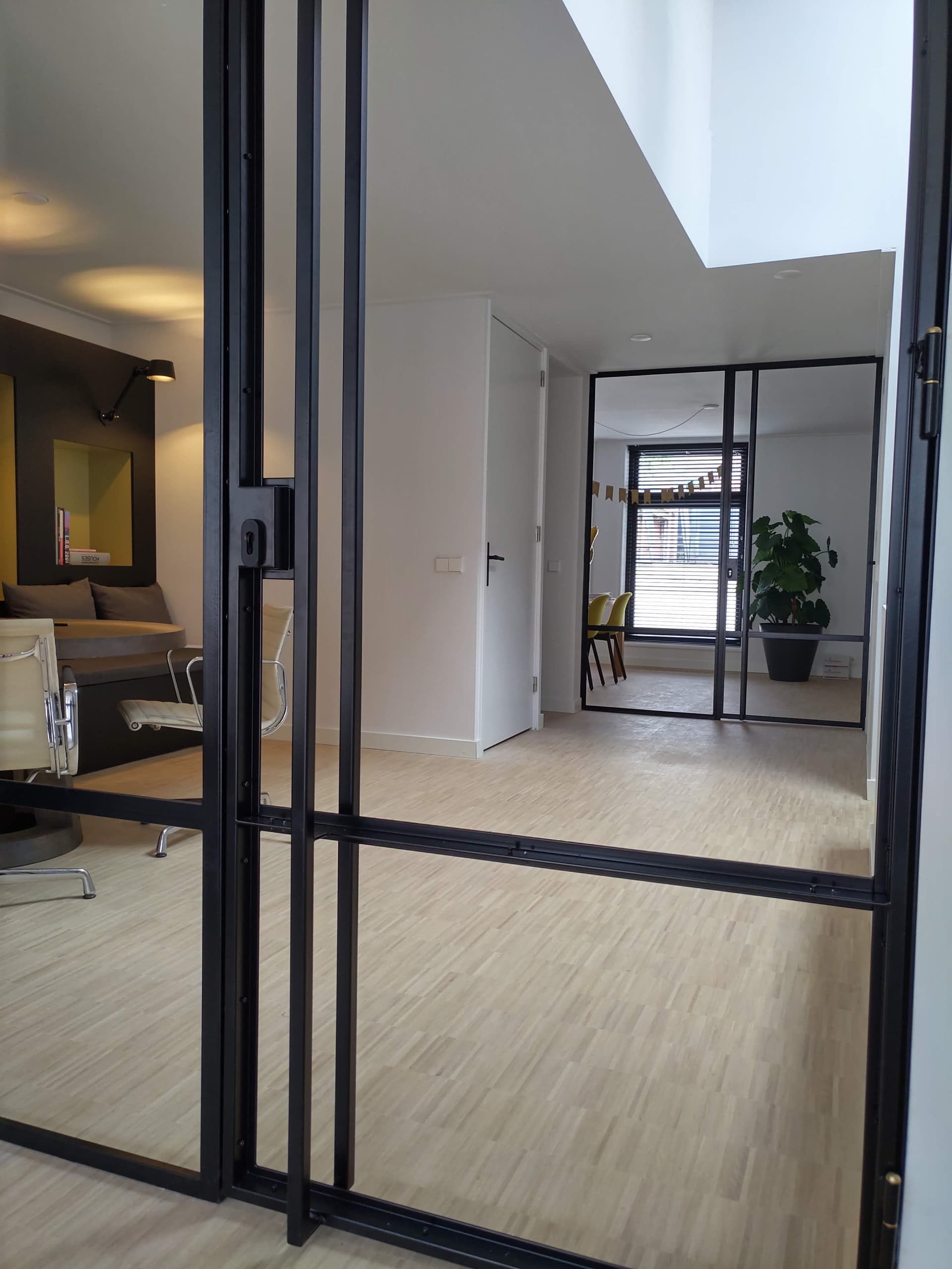 Zwarte stalen binnenpui met scharnierdeur tussen een woonkamer en een hal, met een kamerplant bij het raam erachter.