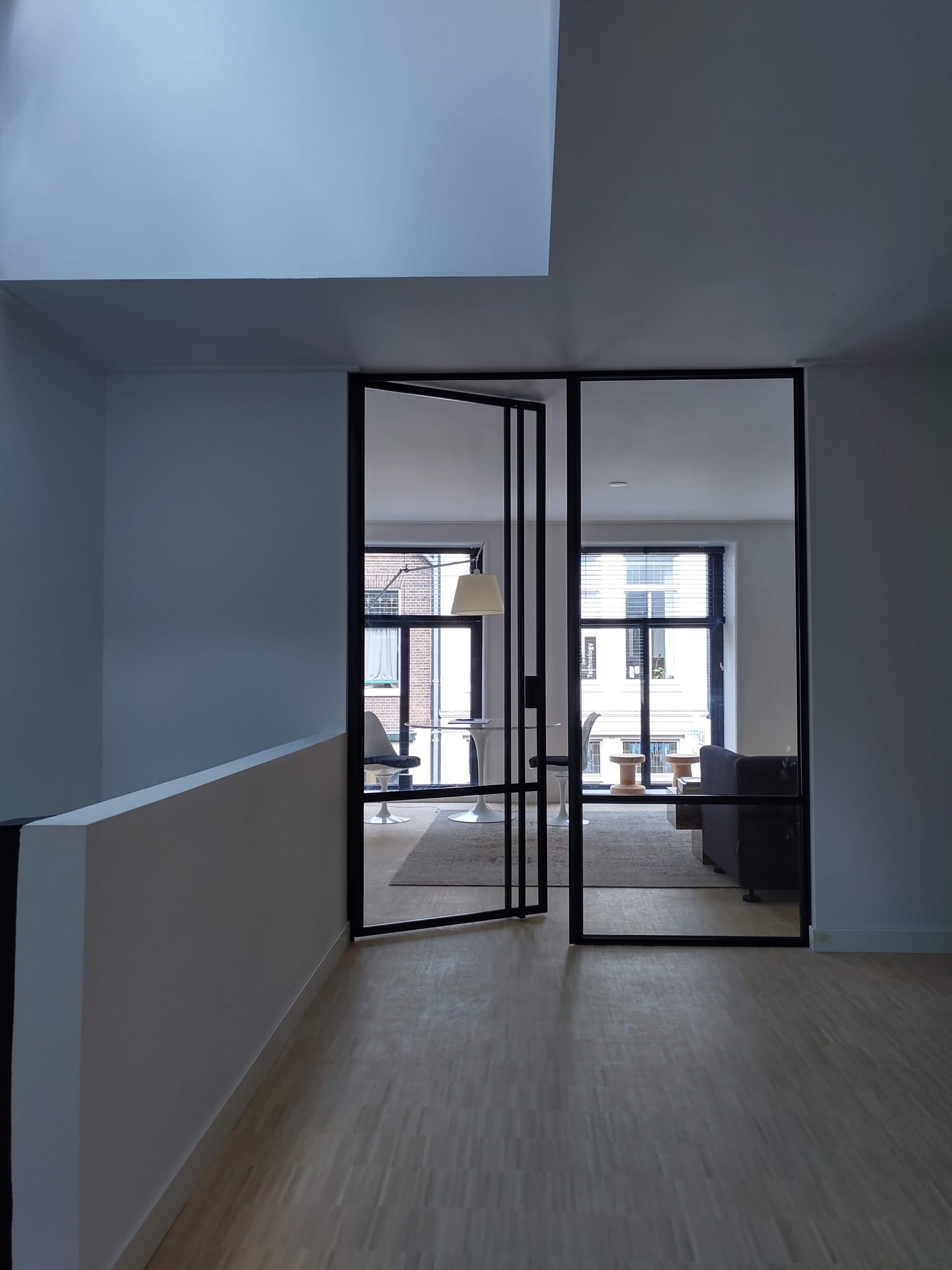 Openstaande zwarte stalen taatsdeur met helder glas tussen een overloop en een lichte werkkamer met raam op de straat.