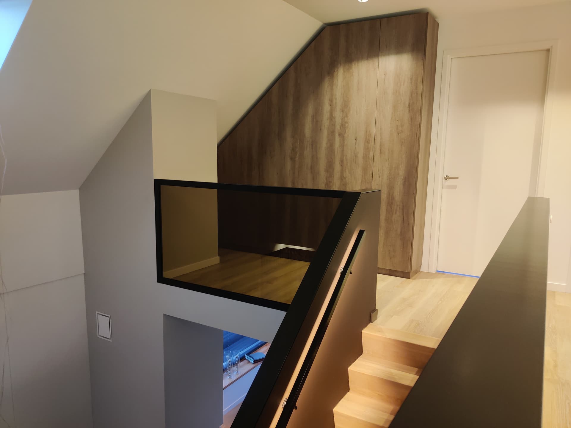 Zwarte stalen balustrade met getint glas langs een vide op de overloop, naast een houten kastenwand en een trap met eikenhouten treden.