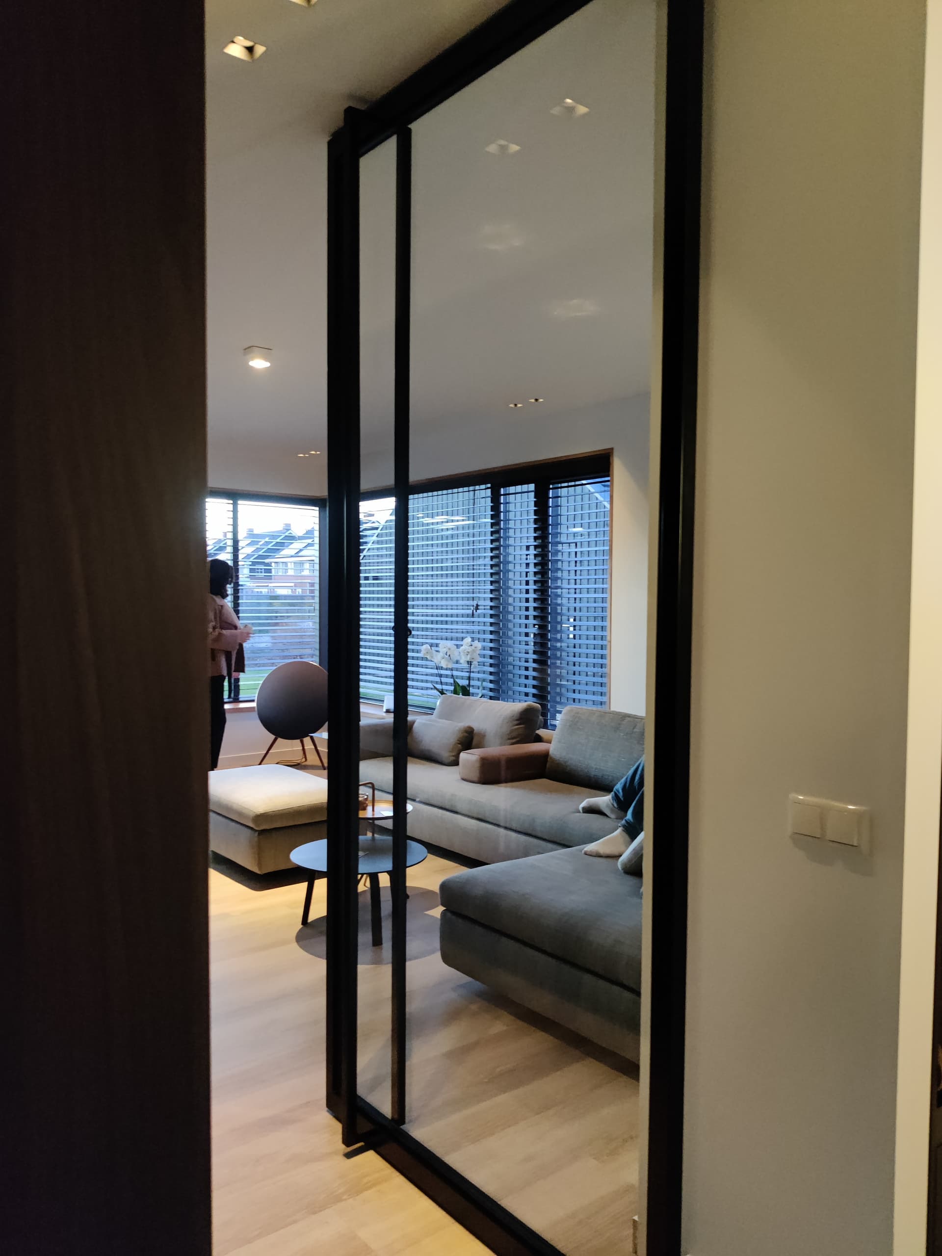 Openstaande minimalistische zwarte stalen taatsdeur, met zicht op een woonkamer met loungebank bij grote ramen.