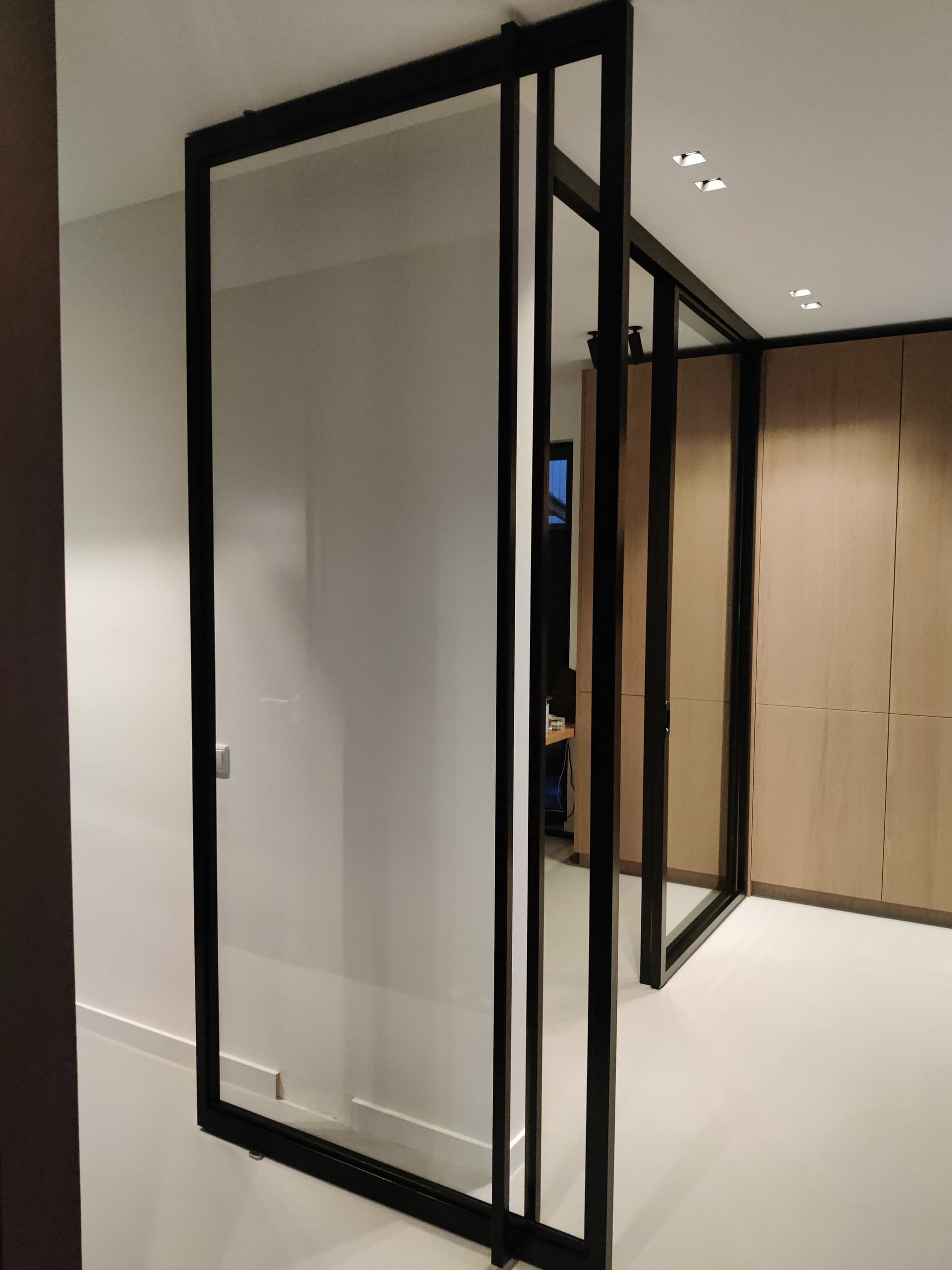 Openstaande minimalistische zwarte stalen taatsdeur met matglas, in een gang met houten kastenwand.