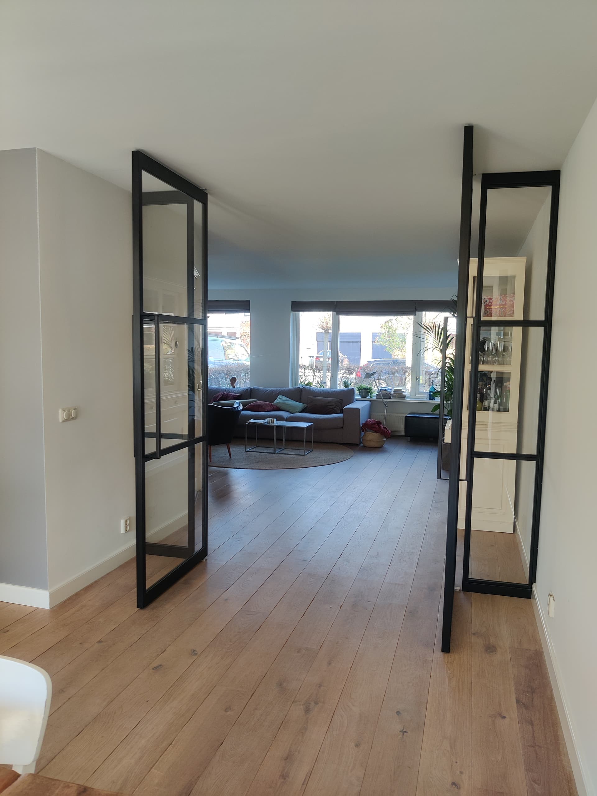 Dubbele zwarte stalen taatsdeuren met zijlichten, half geopend tussen een hal en een woonkamer met houten vloer.