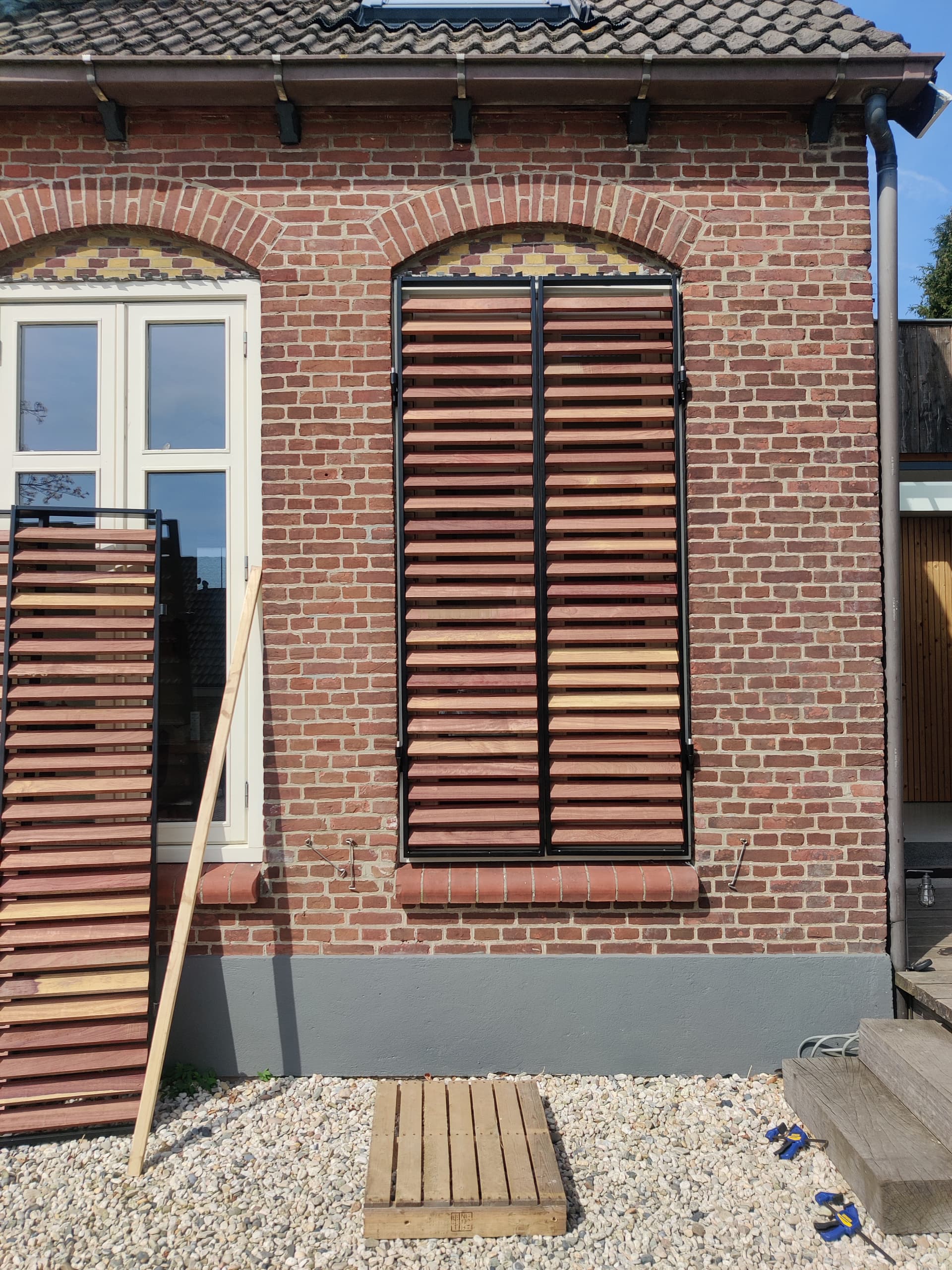 Stalen raamluik met houten lamellen in een boogvenster van een bakstenen gevel, met een tweede luik ernaast klaar om te plaatsen.