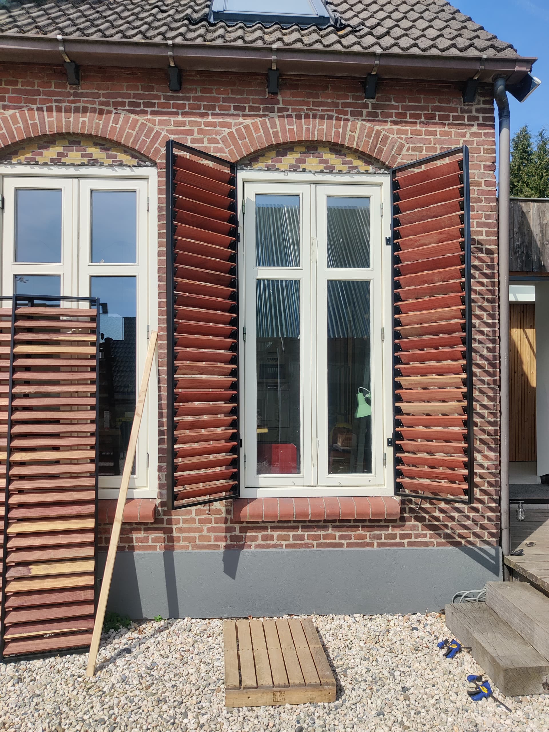 Stalen raamluiken met houten lamellen opengeslagen naast een boogvenster van een bakstenen gevel.