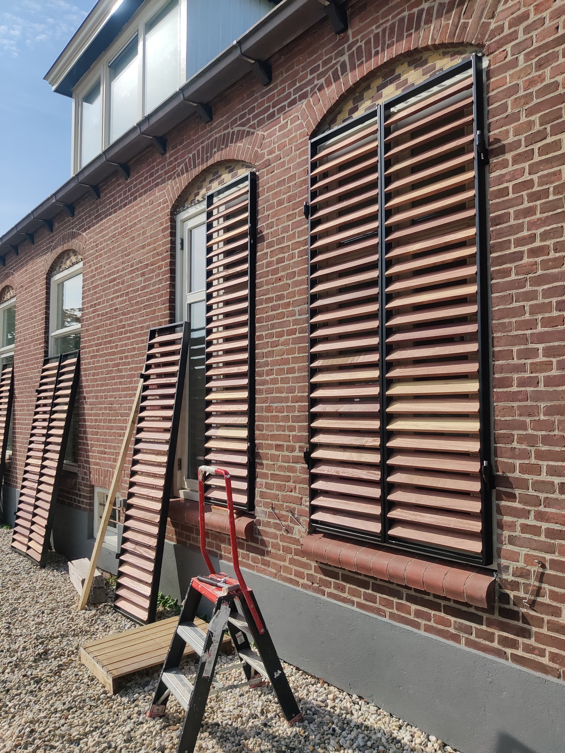 Stalen raamluiken met houten lamellen in de boogramen van een oud bakstenen pand, tijdens de montage.