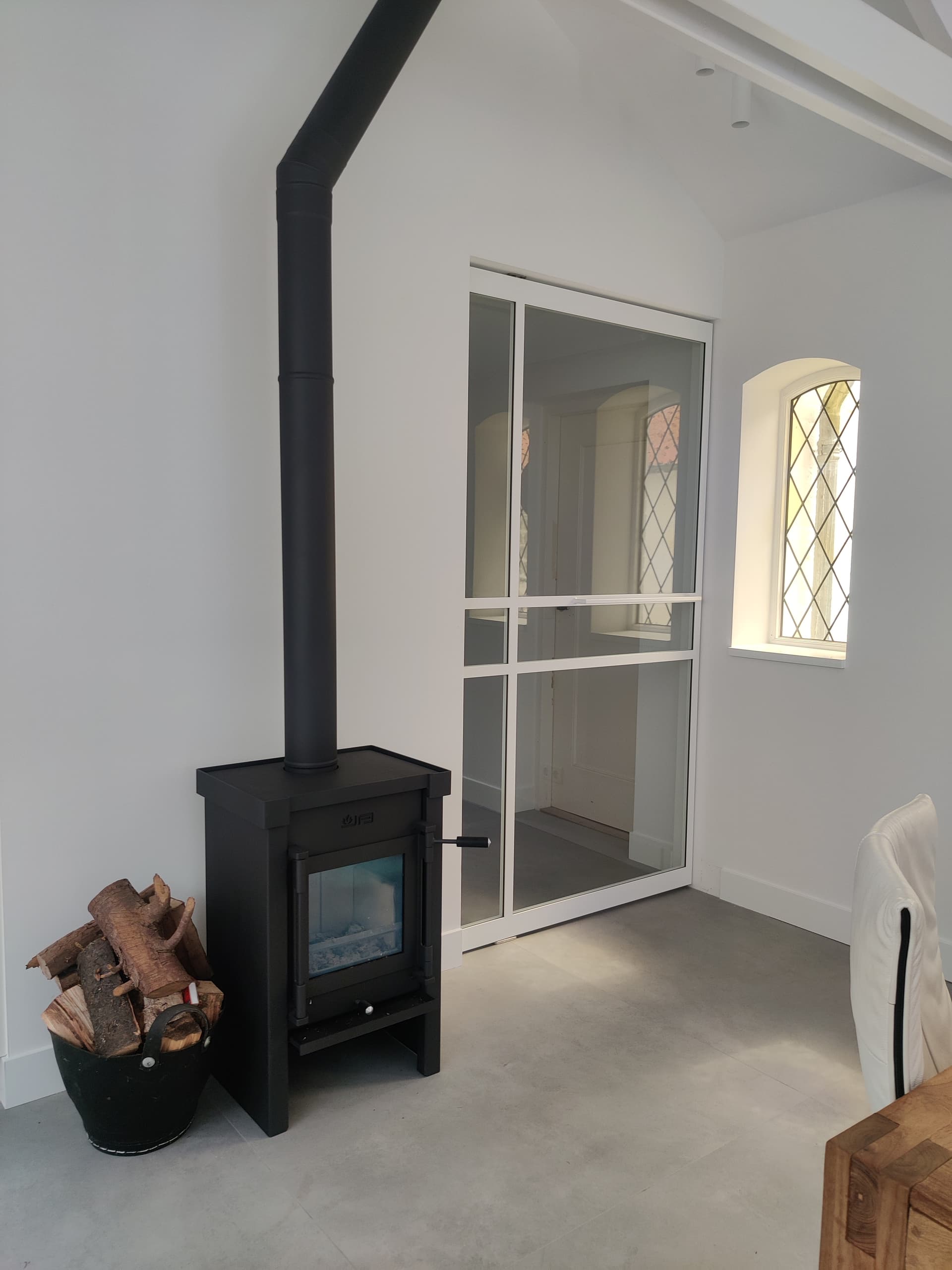 Witte stalen taatsdeur met glas naast een houtkachel, in een ruimte met een glas-in-loodraam.