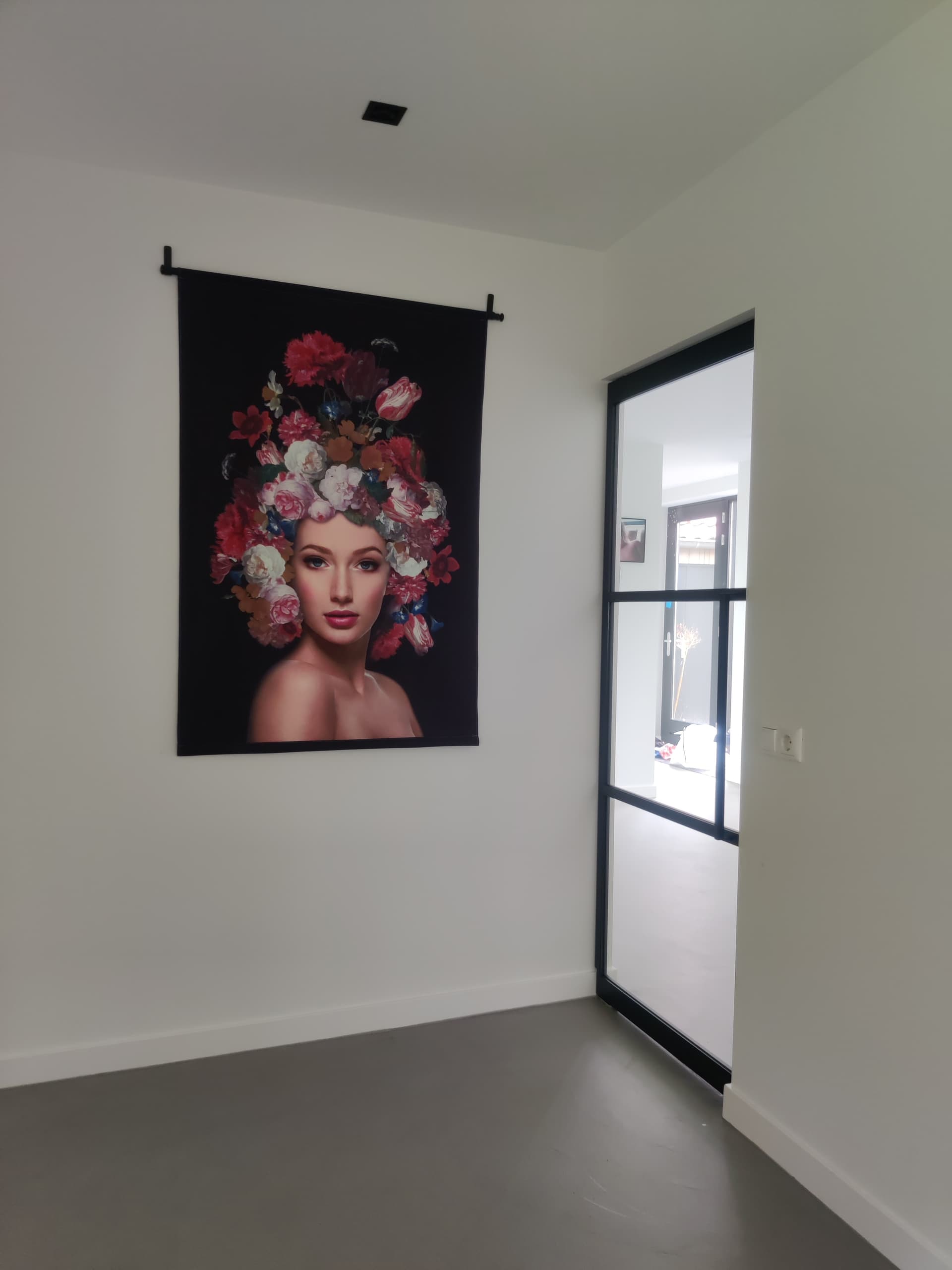 Zwarte stalen taatsdeur met matglas in een witte wand, naast een wandkleed met een portret.
