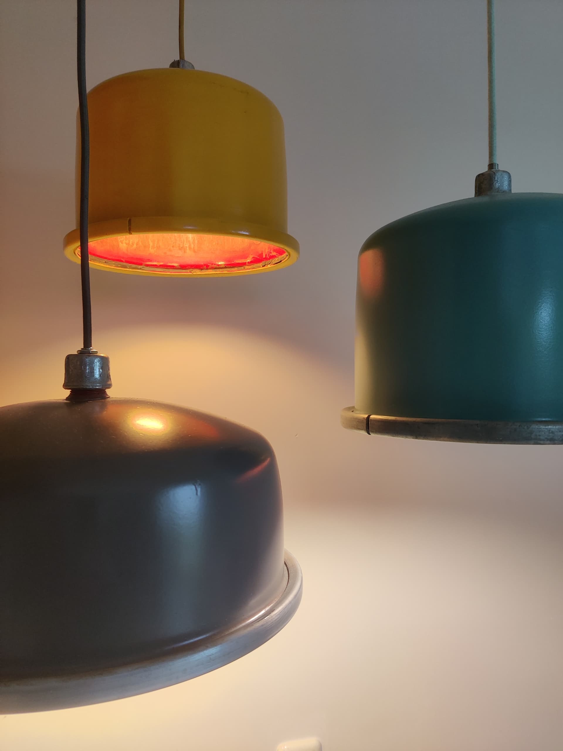 Drie vintage hanglampen in okergeel, petrolblauw en grijs, op verschillende hoogtes opgehangen tegen een witte wand.