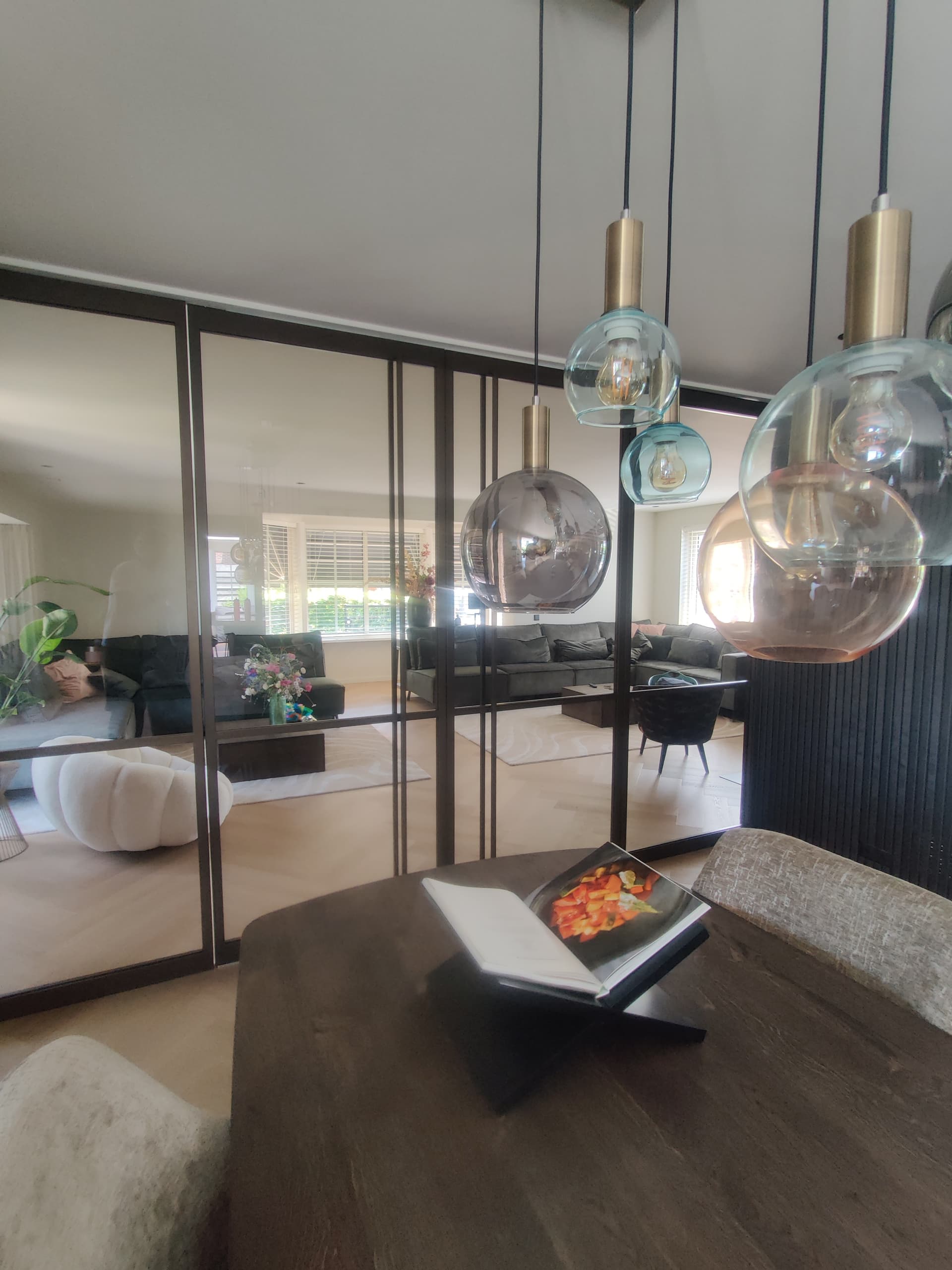 Zwarte stalen binnenpui met dubbele deuren en zijlichten tussen een eetkamer en een woonkamer, met glazen hanglampen ervoor.