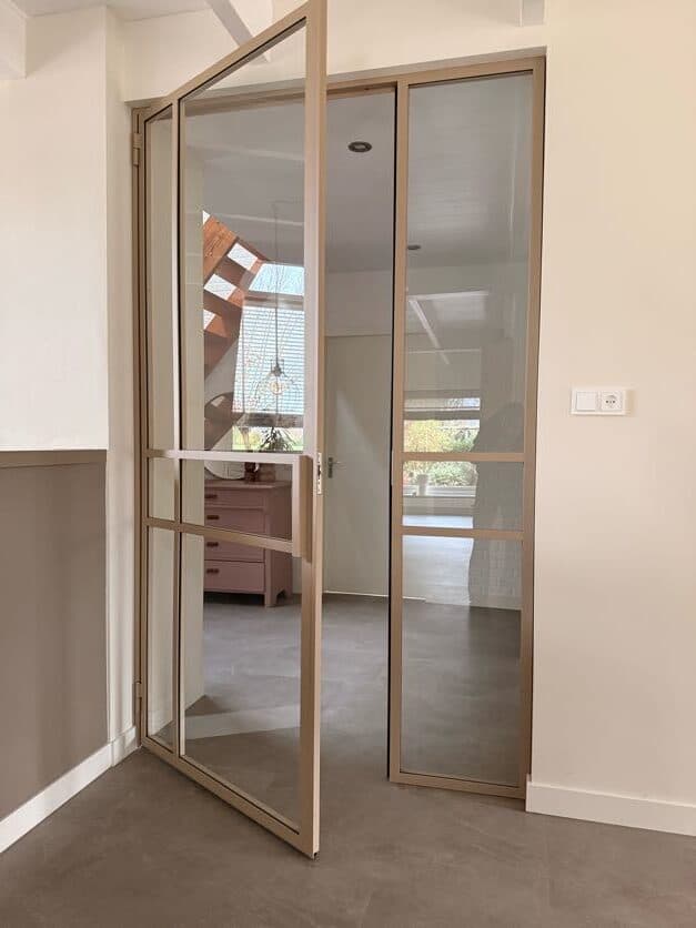 Kamerhoge beige stalen pui met een openstaande scharnierdeur, vast zijpaneel en helder glas gemonteerd in een licht interieur.