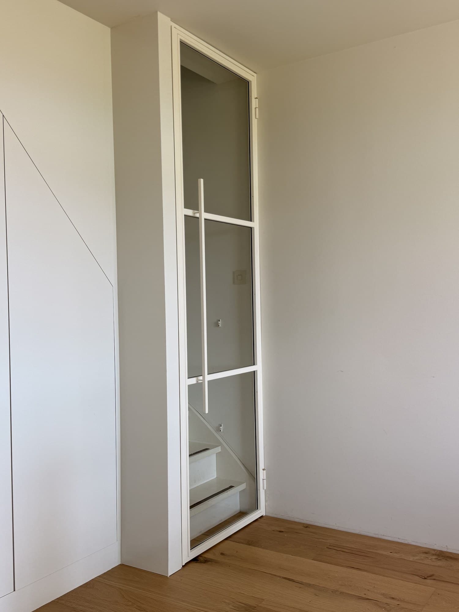 Witte stalen scharnierdeur met drie rechte vakken en glas, gesloten in een lichte hal met eiken vloer, met de trap erachter zichtbaar.