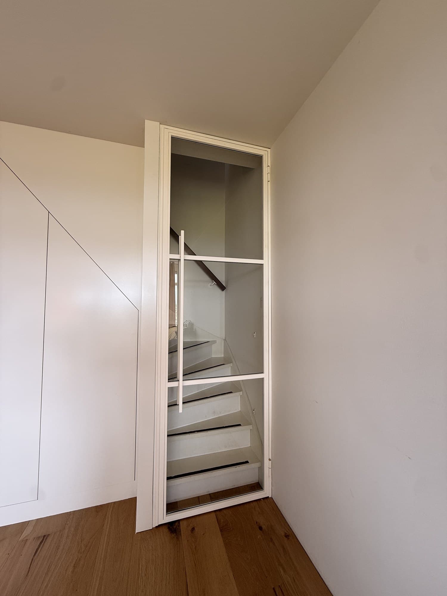 Witte stalen scharnierdeur op een kier, met zicht op de witte trap erachter.