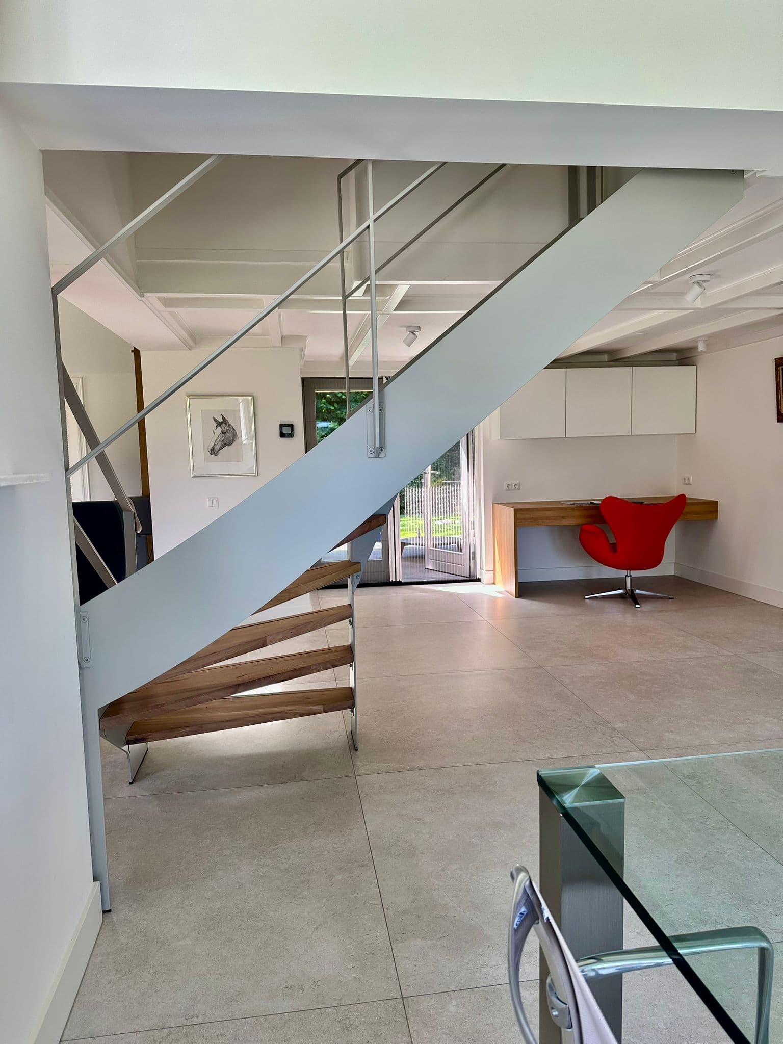 Open stalen designtrap met lichtgrijze coating en eikenhouten treden, met zicht op een werkplek erachter.