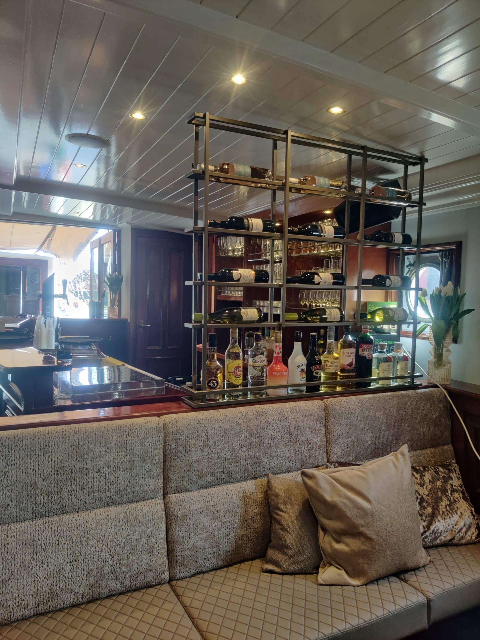 Goudkleurig stalen wijnrek als roomdivider boven een bank, met flessen en glazen, in de salon van een schip.