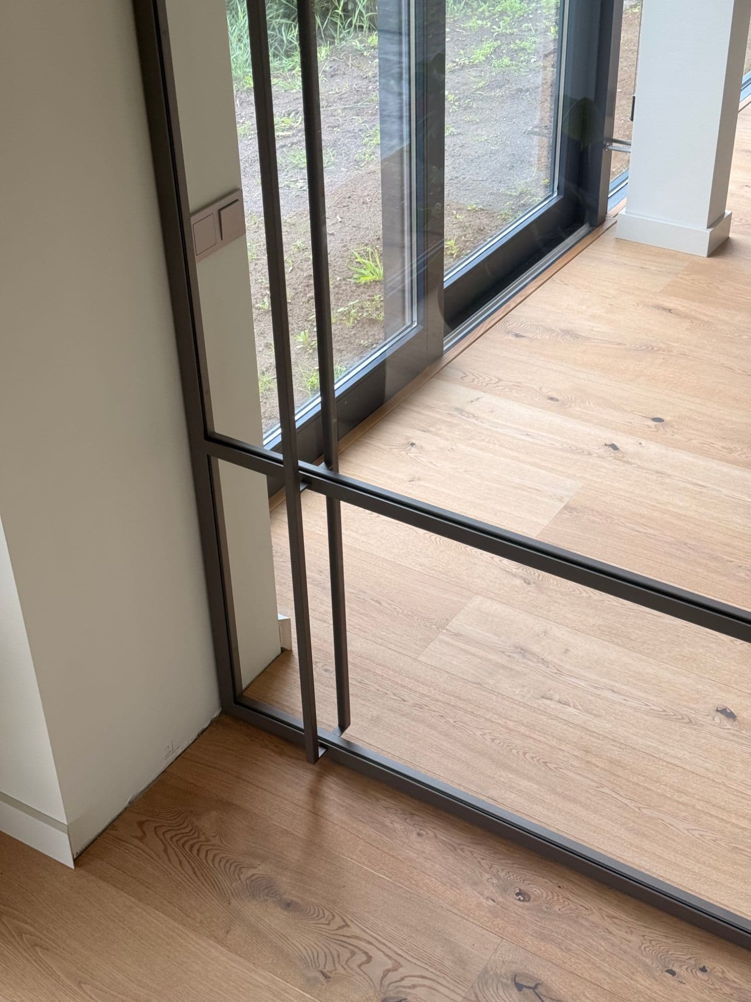Detail van de bruine stalen schuifdeur van bovenaf, met de dubbele verticale stijl die als greep dient en de dwarsregel boven de eiken vloer.