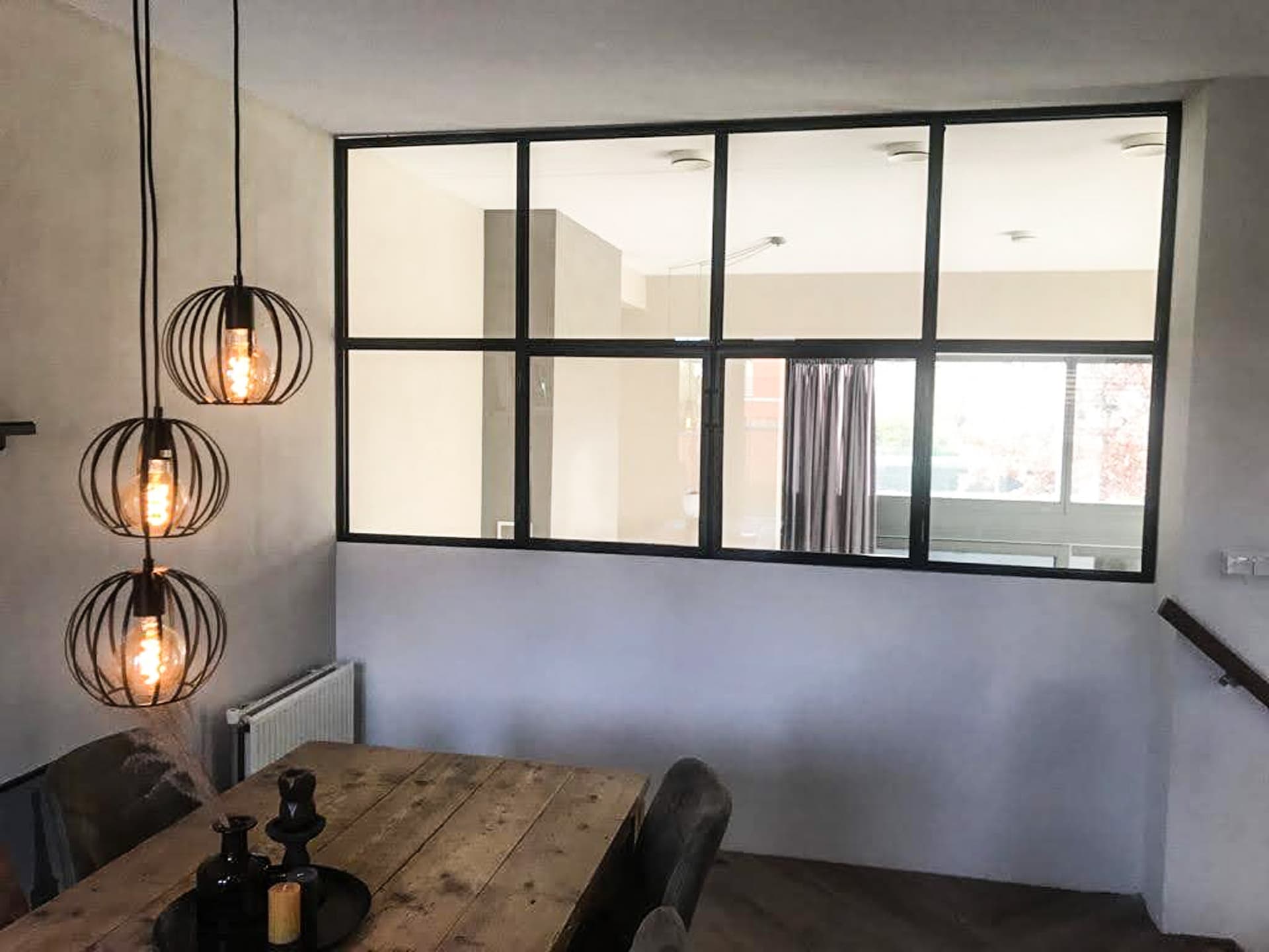 Zwart stalen binnenraam met zesdelige roedeverdeling in een betonlook wand, boven een houten eettafel met hanglampen.