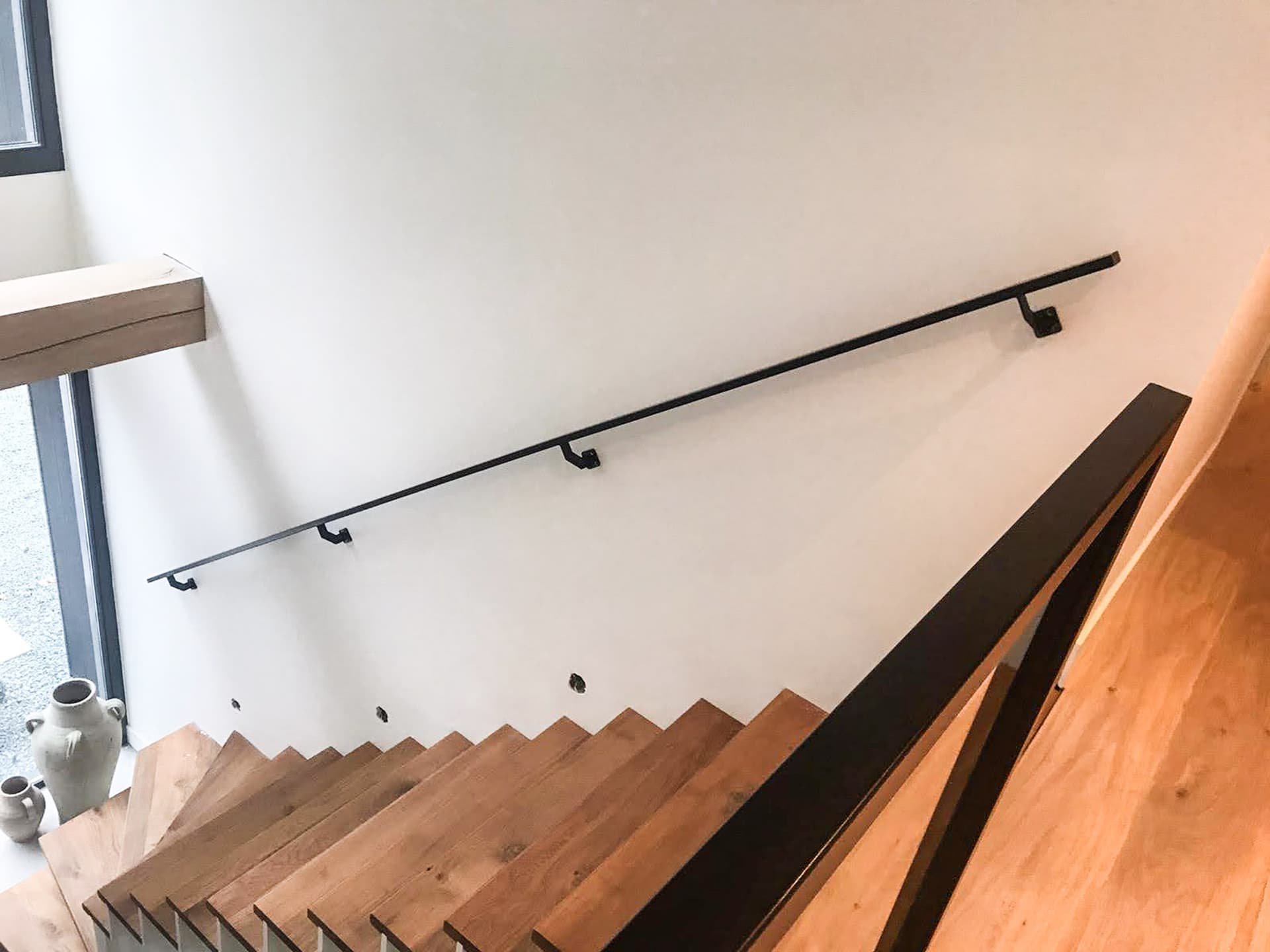 Zwarte stalen muurleuning met beugels langs een open trap, met aan de overzijde een stalen balustrade van dezelfde lijn.