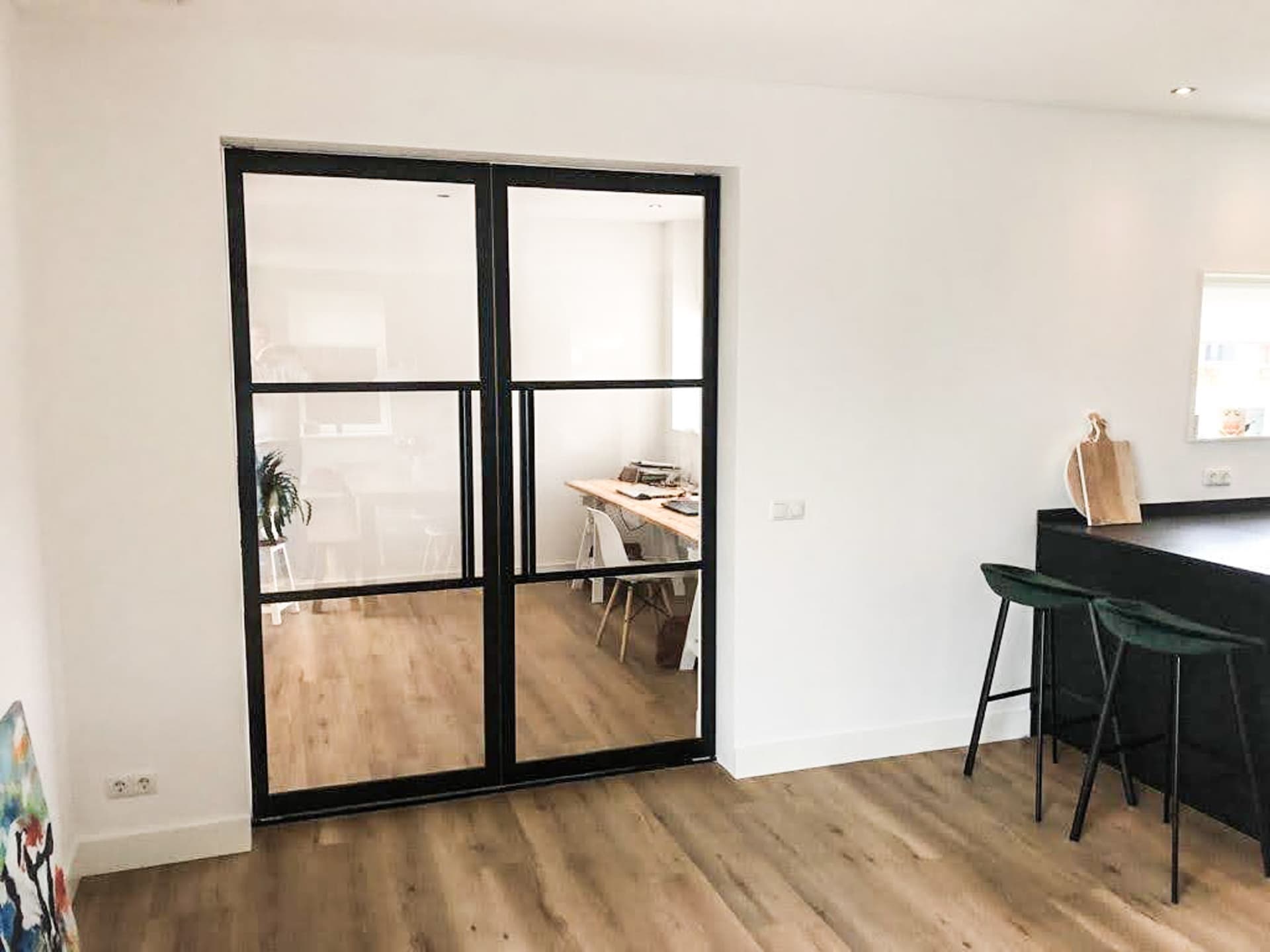Dubbele zwarte stalen taatsdeuren met heldere beglazing tussen een woonkeuken en een werkkamer met eettafel.