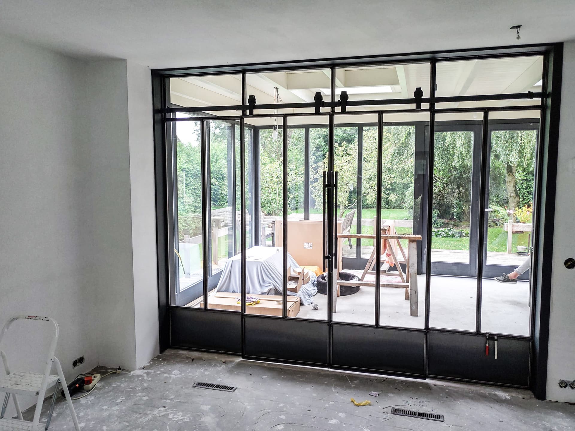 Zwarte stalen pui met schuifdeuren en bovenlichten tussen een woonkamer in aanbouw en de tuin.