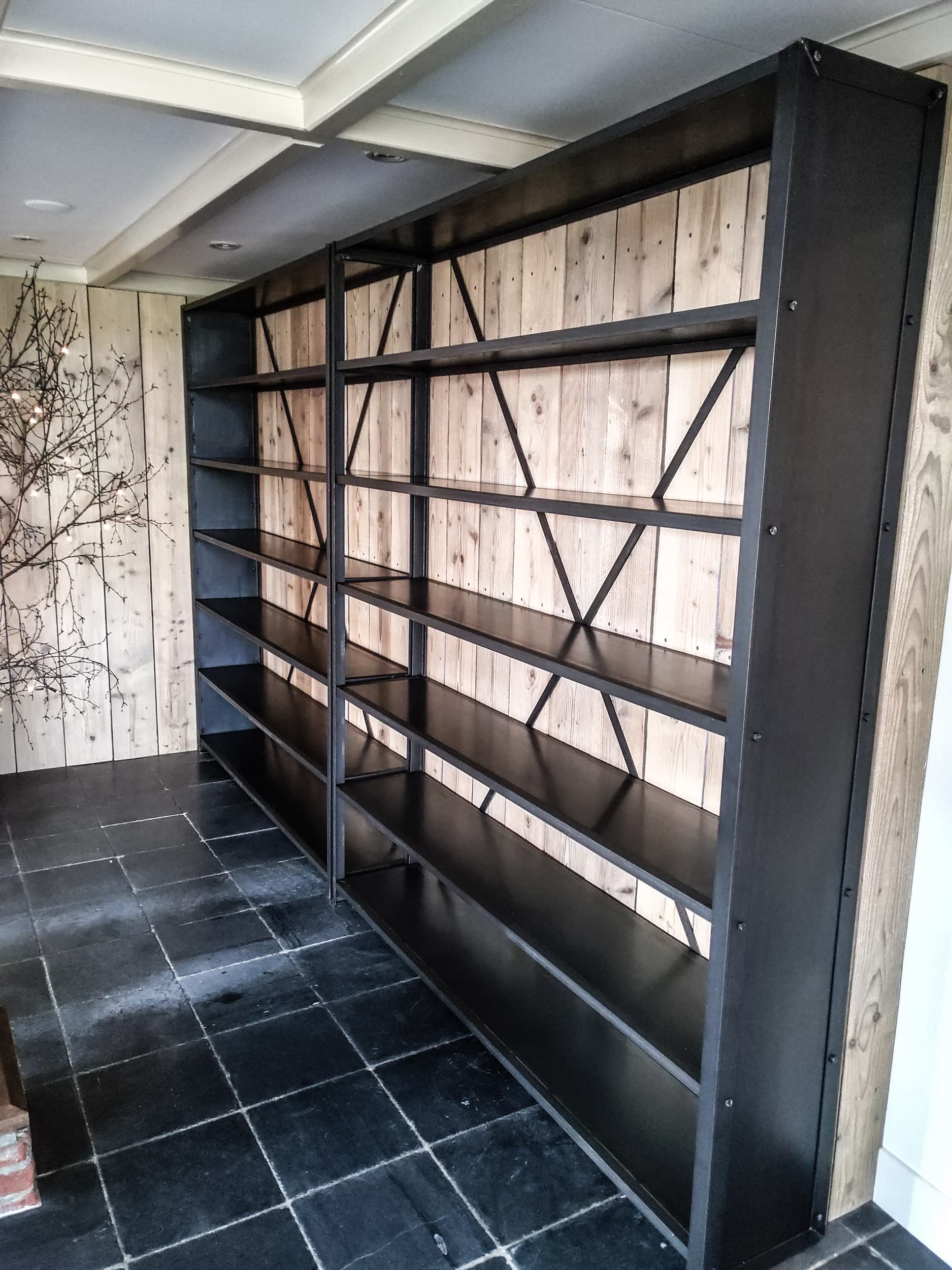 Grote zwarte stalen stellingkast van vloer tot plafond met kruisverbanden, tegen een houten wand boven een donkere plavuizenvloer.
