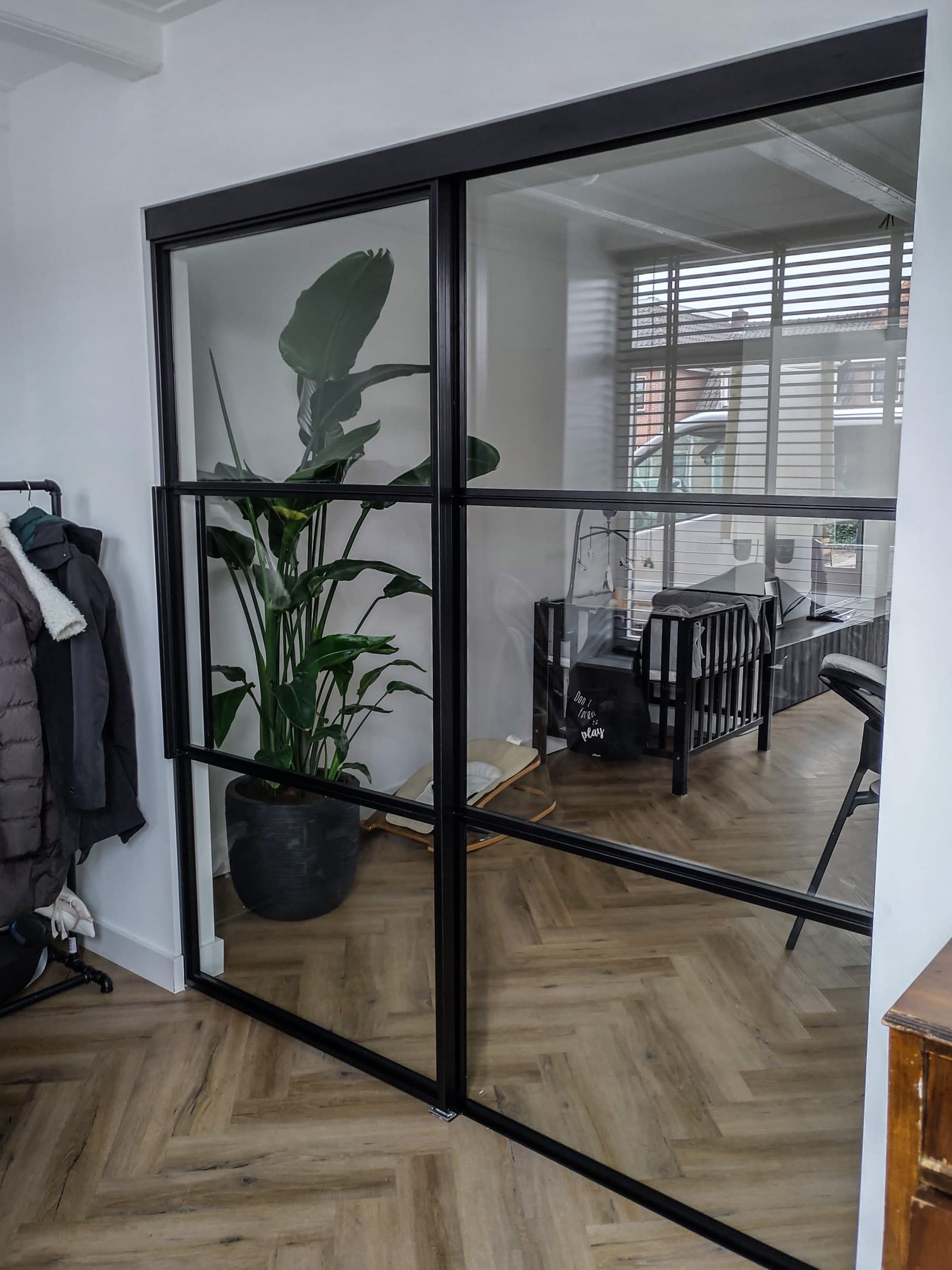 Zwarte stalen binnenpui met dubbele deuren en heldere beglazing in een hal met visgraatvloer, met zicht op een kamerplant en de woonkamer.