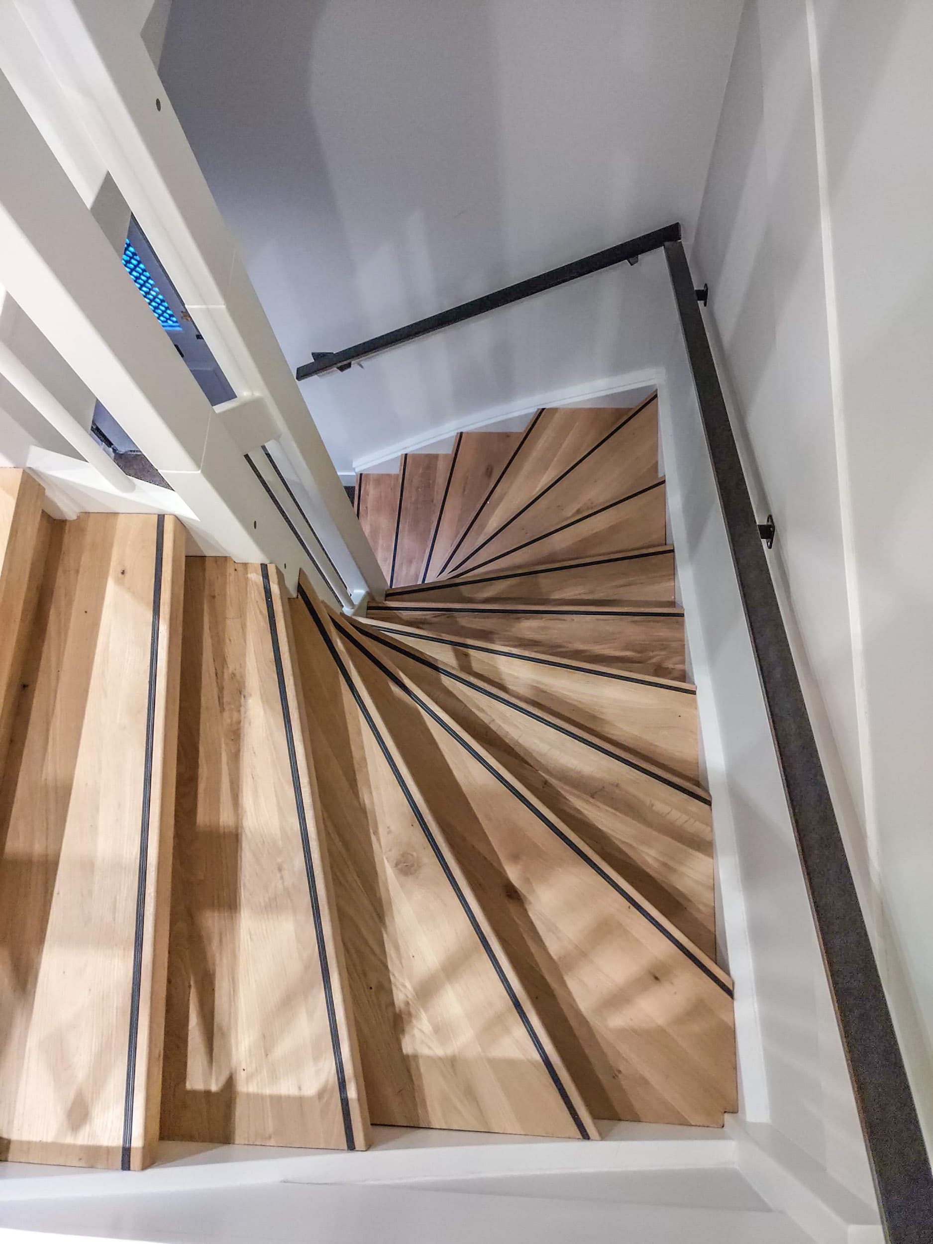 Slanke zwarte stalen trapleuning langs een houten kwartslagtrap, van bovenaf gezien in een wit trappenhuis.