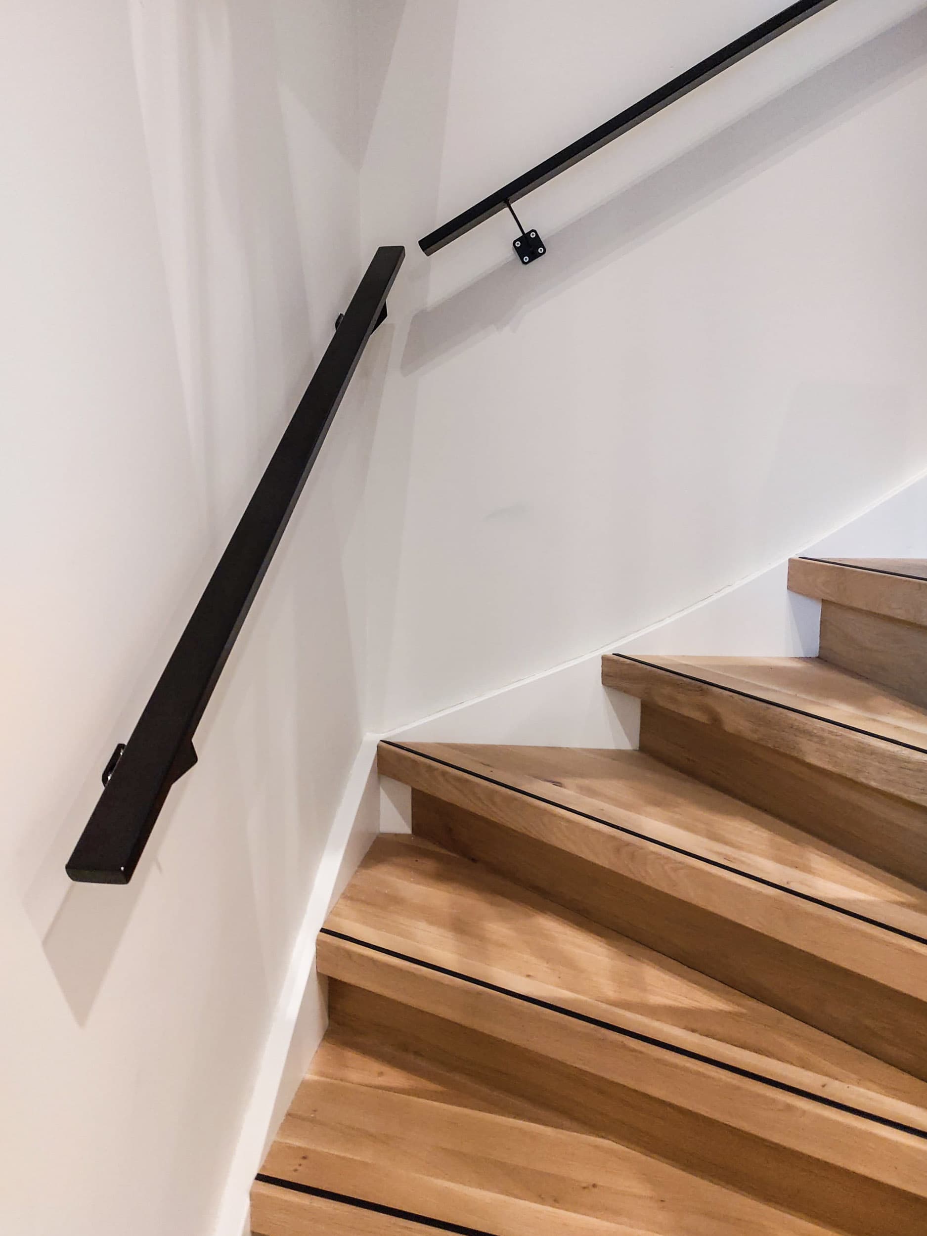 Zwarte stalen muurleuning met zichtbare bevestigingsbeugels langs een eikenhouten trap in een wit trappenhuis.