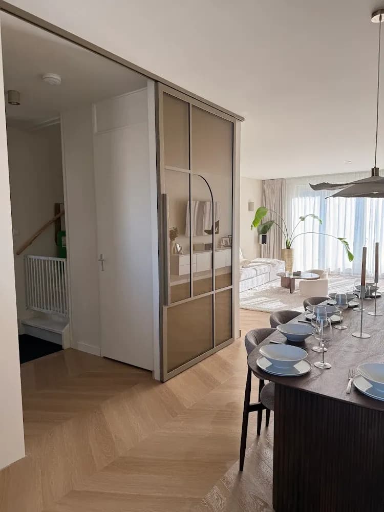 Luxe openstaande bronzen stalen binnendeur op rails in een sfeervolle woonkamer met visgraatvloer en houten eettafel.