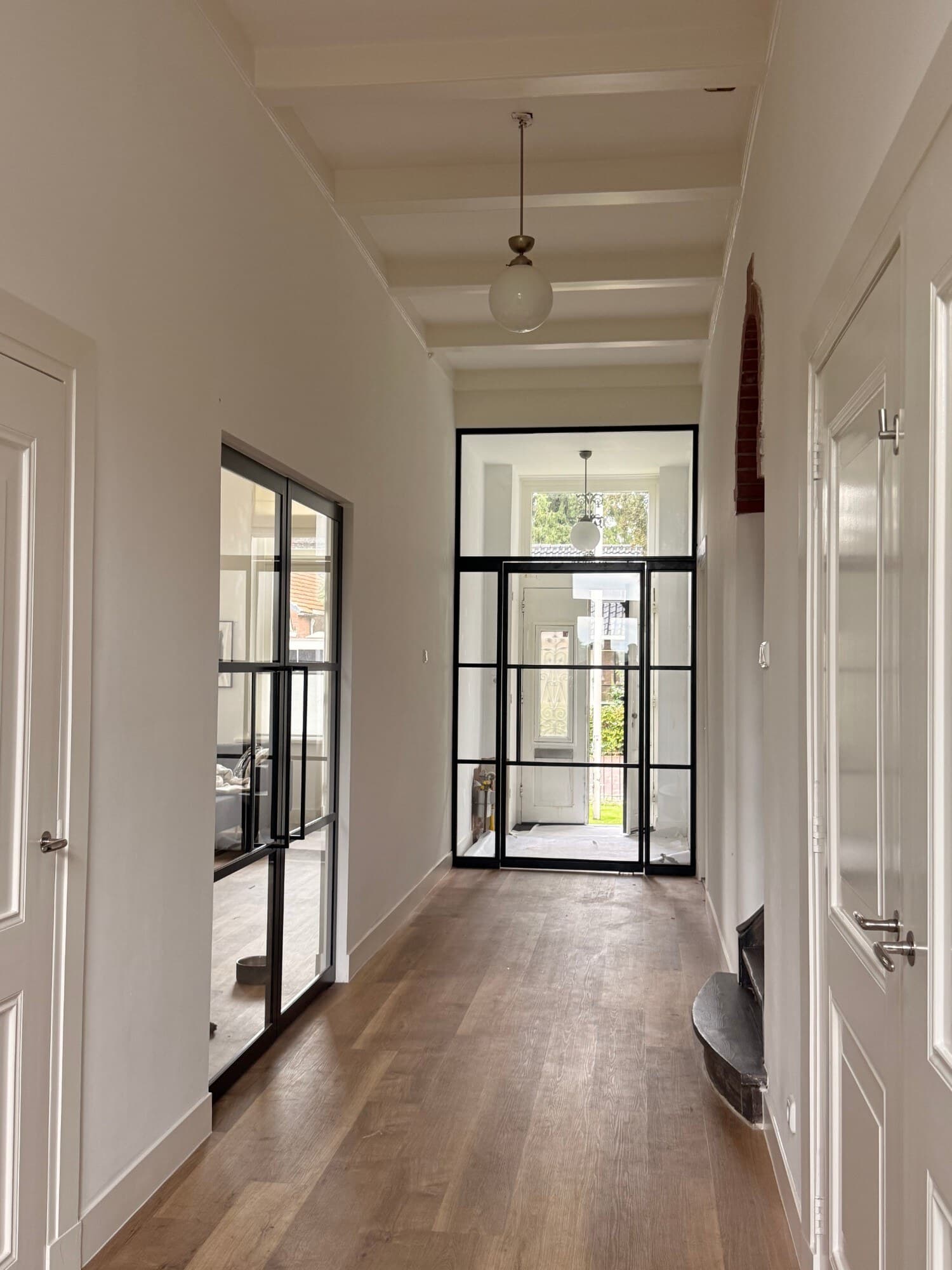 Lange gang met balkenplafond en houten vloer, met aan het eind de zwarte stalen taatsdeur met zijlichten en bovenlicht naar het portaal.