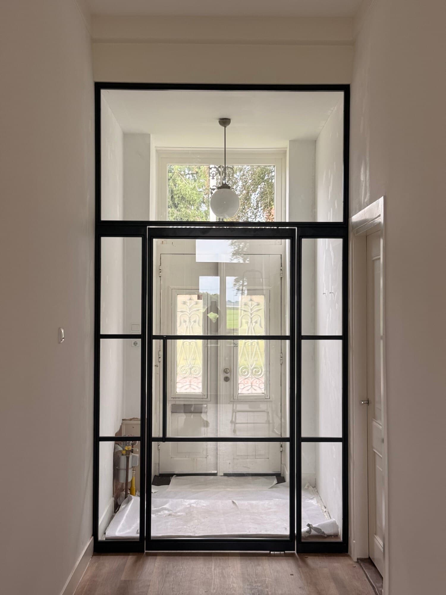 Kamerhoge zwarte stalen taatsdeur met een zijlicht links en rechts en een groot bovenlicht, gesloten, met door het heldere glas de oude voordeur van het portaal.