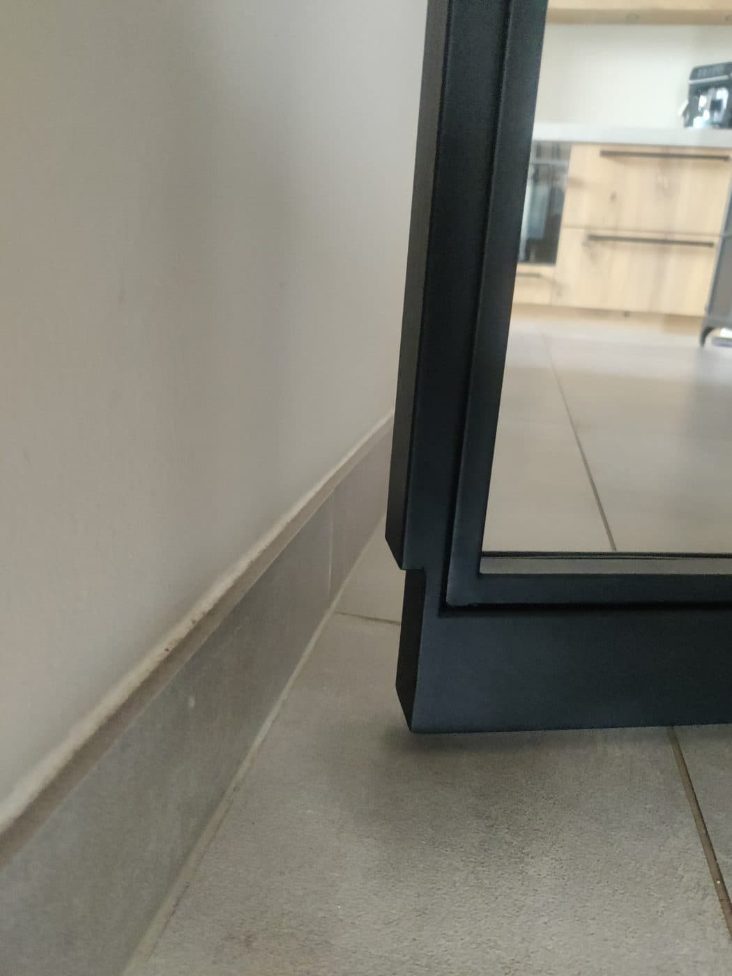 Onderhoek van een zwart stalen taatsdeurkozijn op een tegelvloer, waarbij de vloerplaat bovenop de tegels ligt in plaats van erin verzonken.