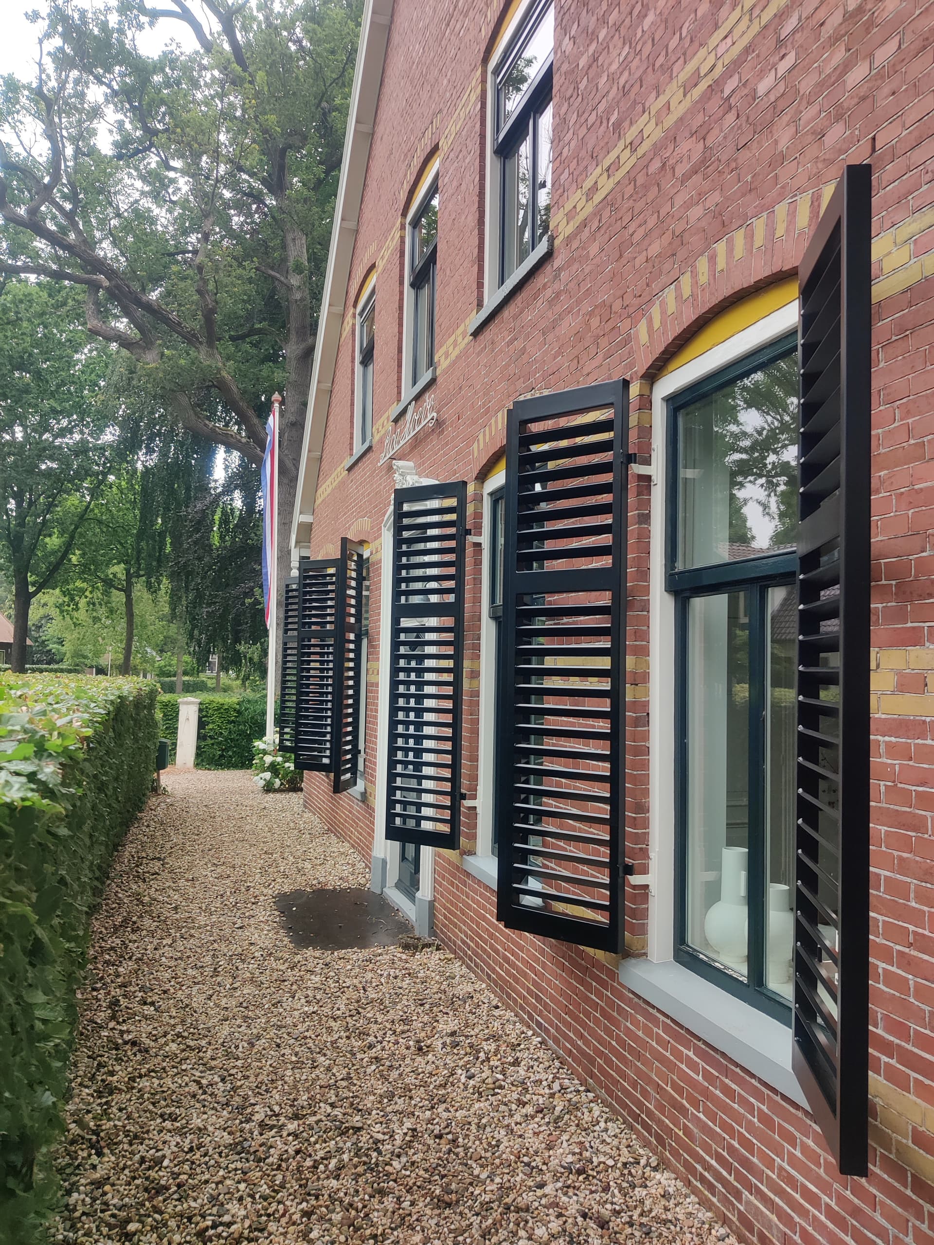 Zwarte stalen luiken met lamellen langs de ramen van een oude boerderij van rode baksteen, met een grindpad langs de gevel.