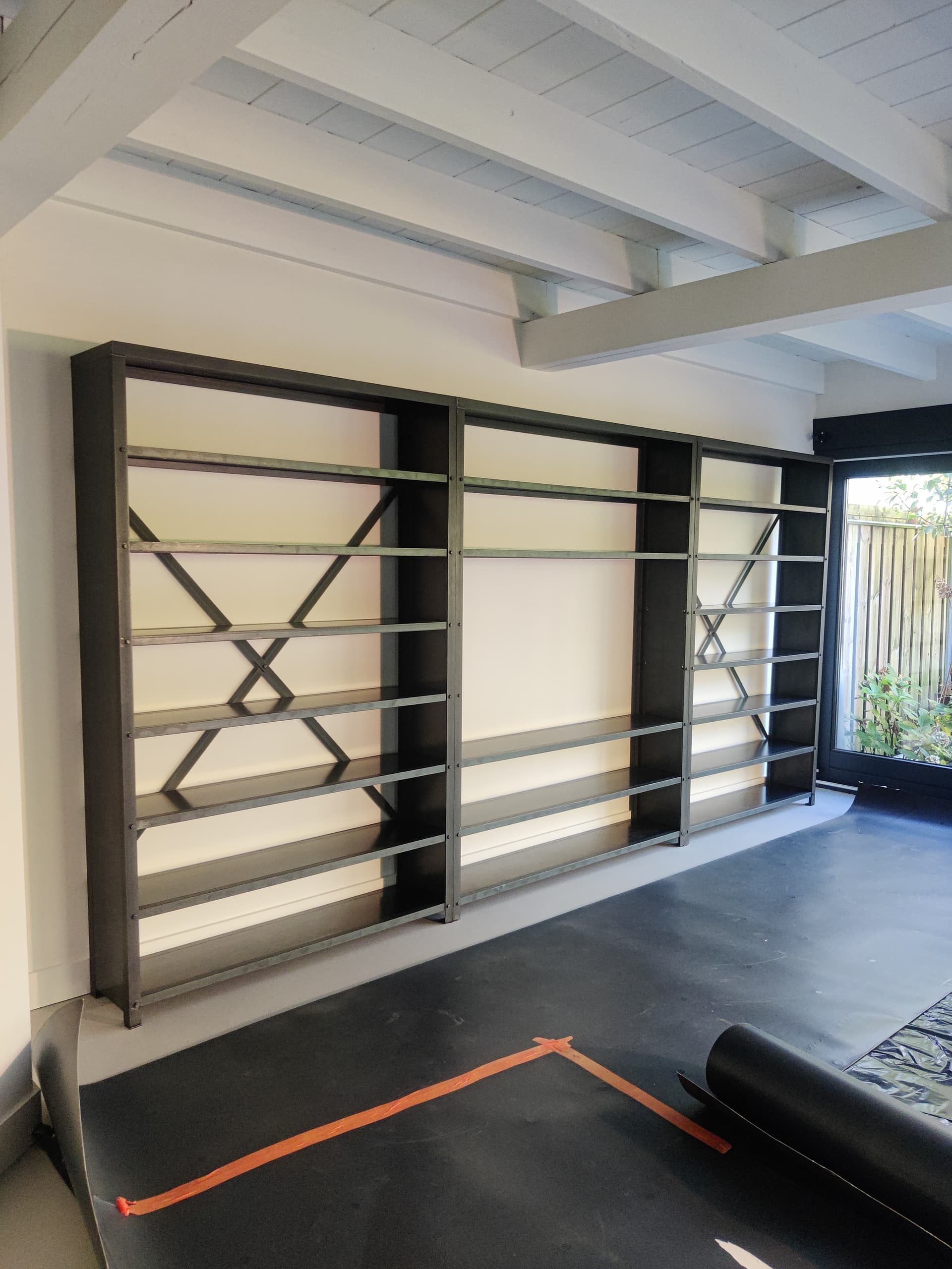 Drie stalen wandkasten in blank staal met kruisverbindingen, over de volle breedte van een witte wand onder een balkenplafond.