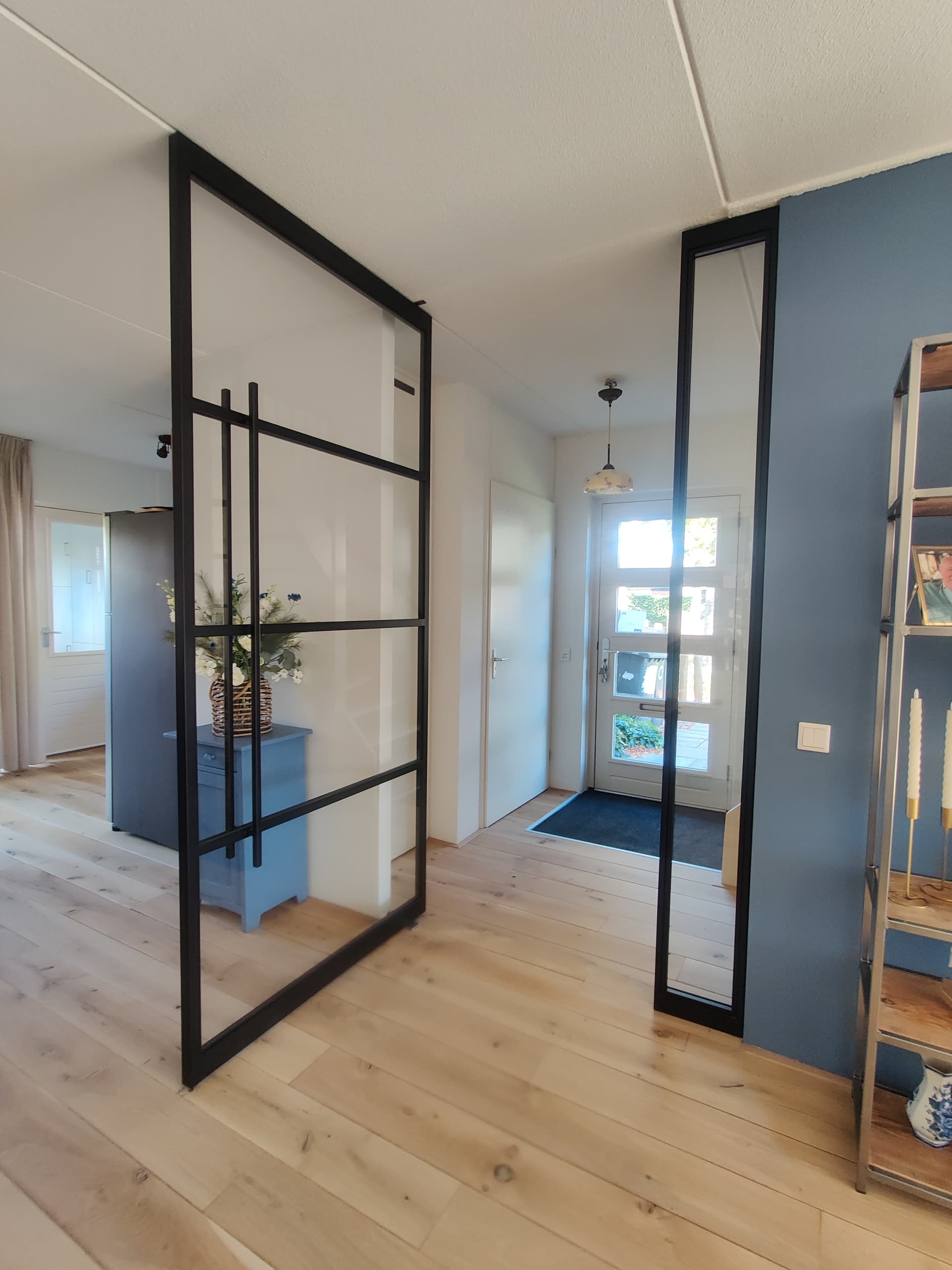 Stalen taatsdeur met helder glas, opengedraaid om zijn as in een hal, naast een smal vast zijlicht in een blauwe wand.